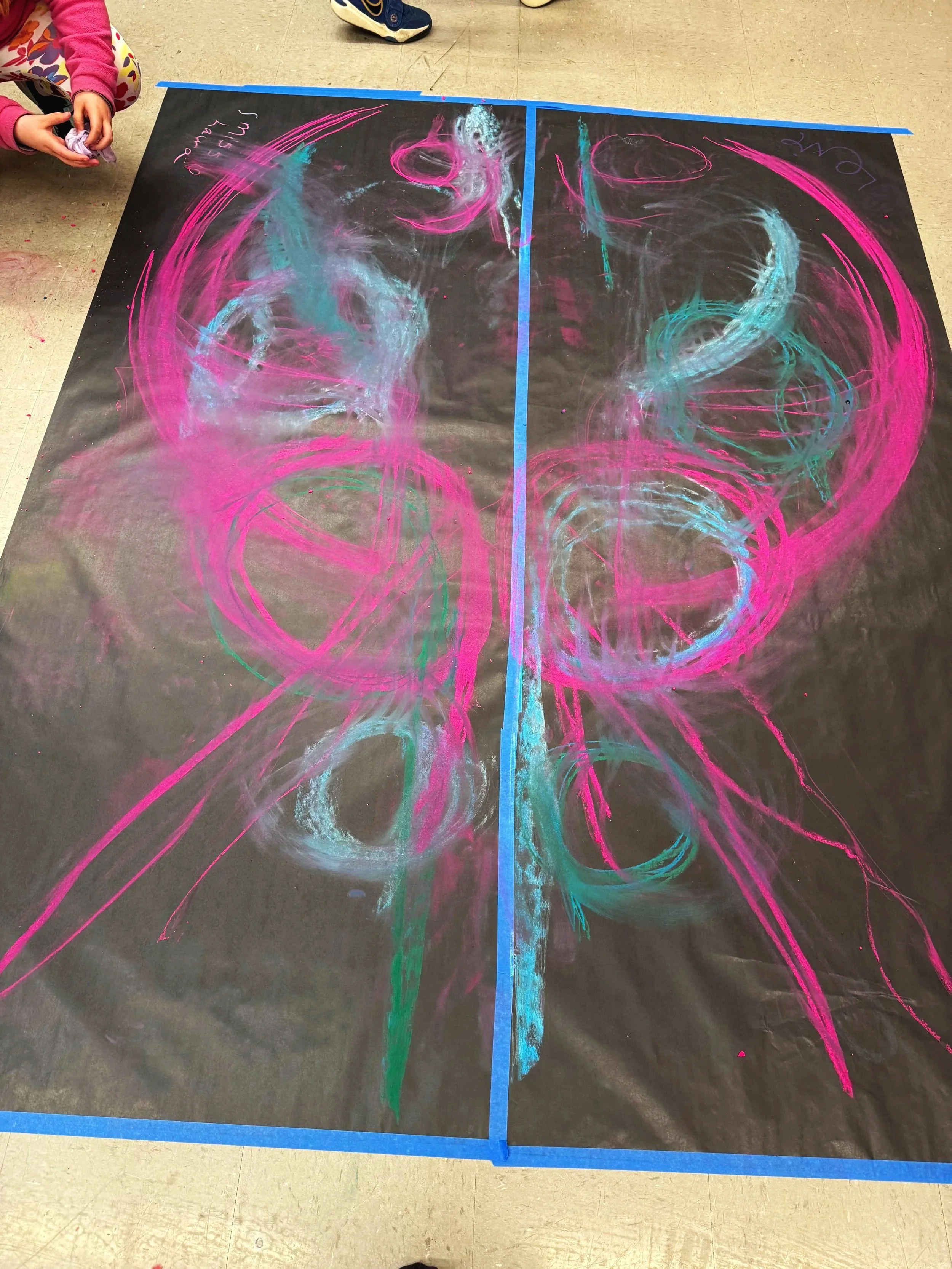 Child's hand holding pink chalk, colorful abstract chalk drawing on large black paper floor