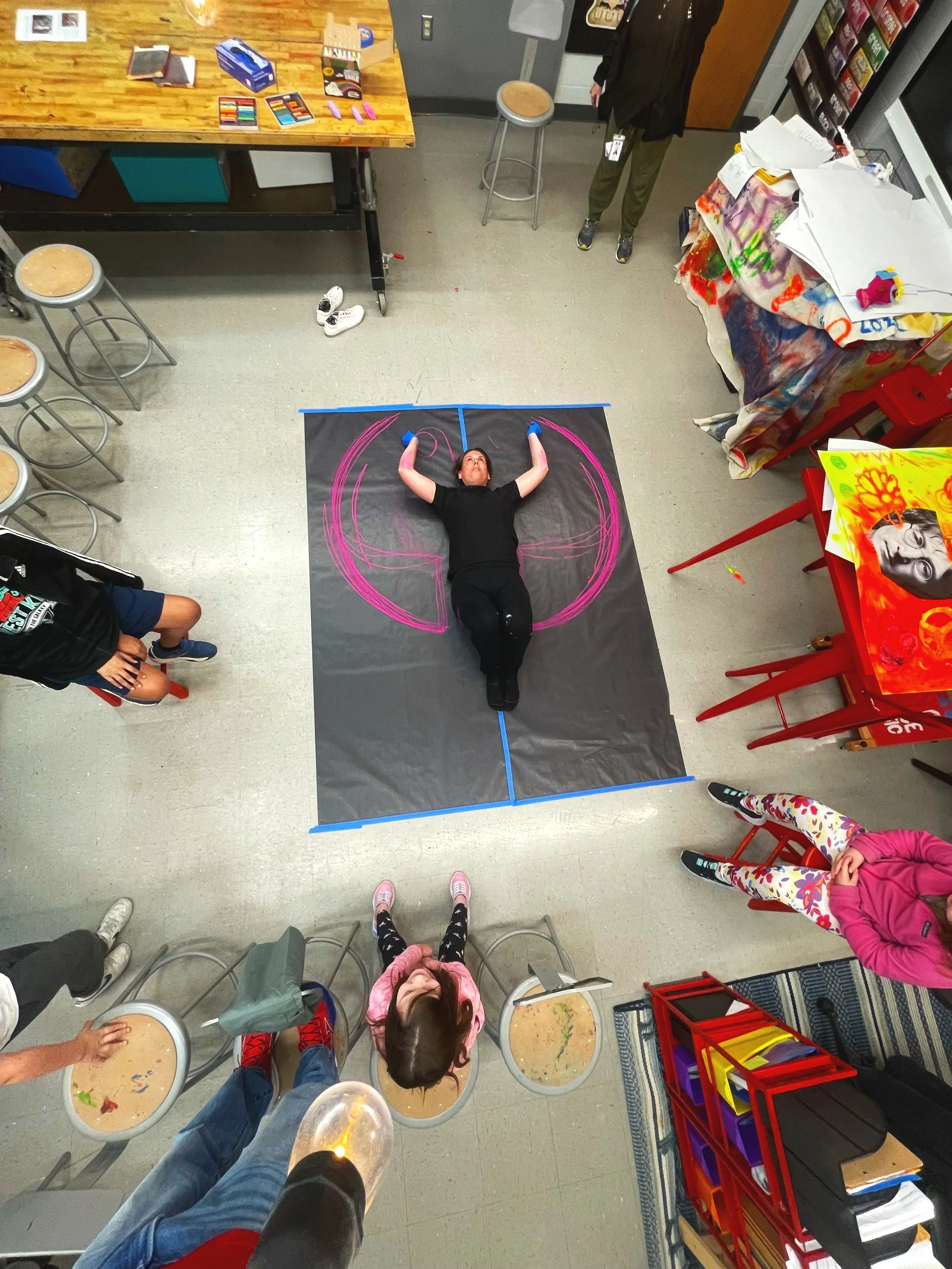 A woman lying on a black mat in an art classroom with pink chalk markings on the floor around her, seen from above, with children and art supplies surrounding her.