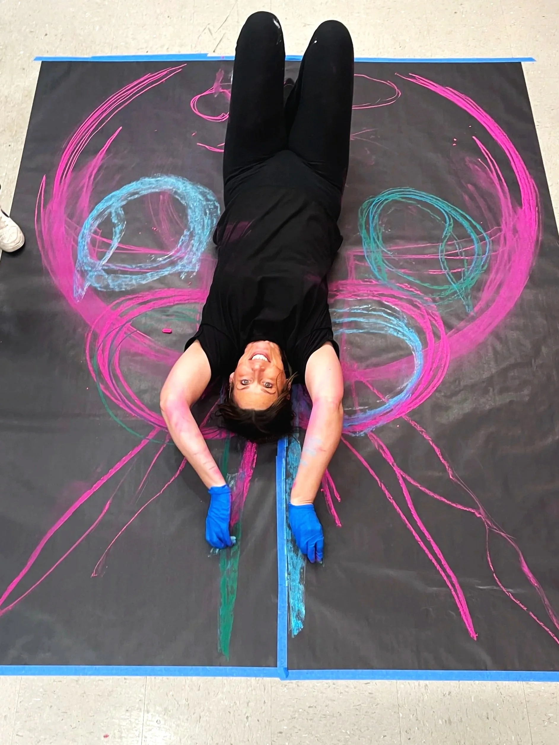 A woman lying on a large black canvas with colorful chalk drawings of abstract pink, blue, and green shapes, smiling, wearing black clothes and blue gloves.