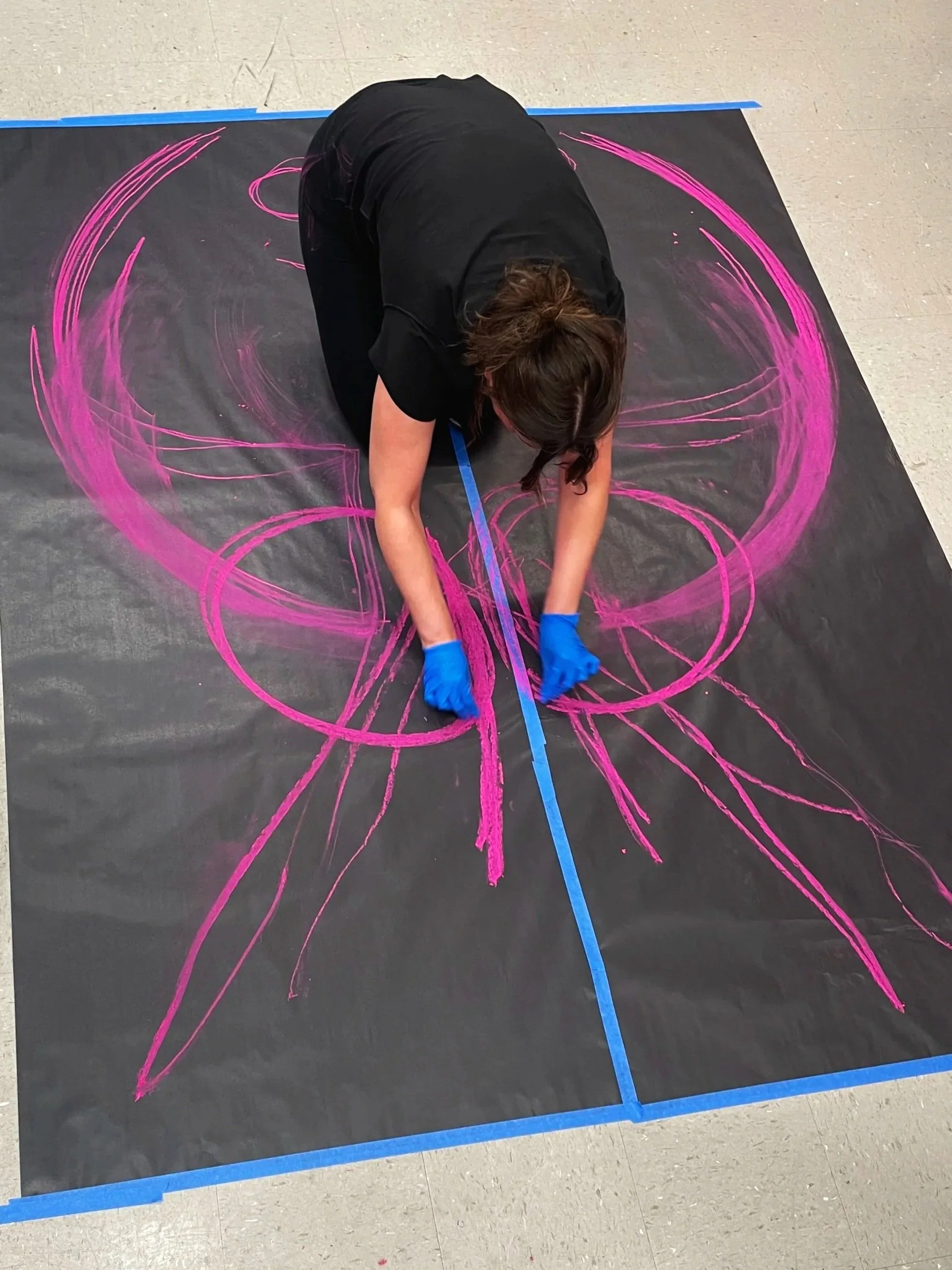 An artist creates vibrant pink chalk art on a large black surface, using tape to guide their intricate design.