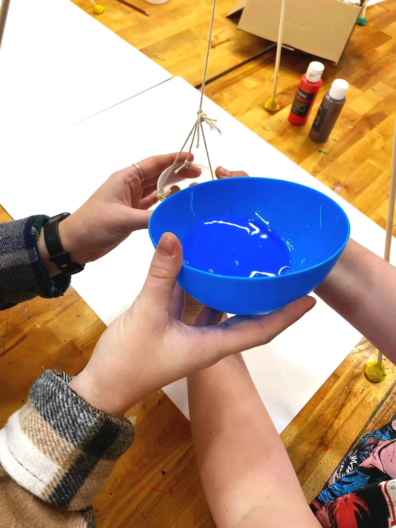 Two people working together on a science experiment involving a blue plastic bowl and a suspension setup with strings, on a wooden table.