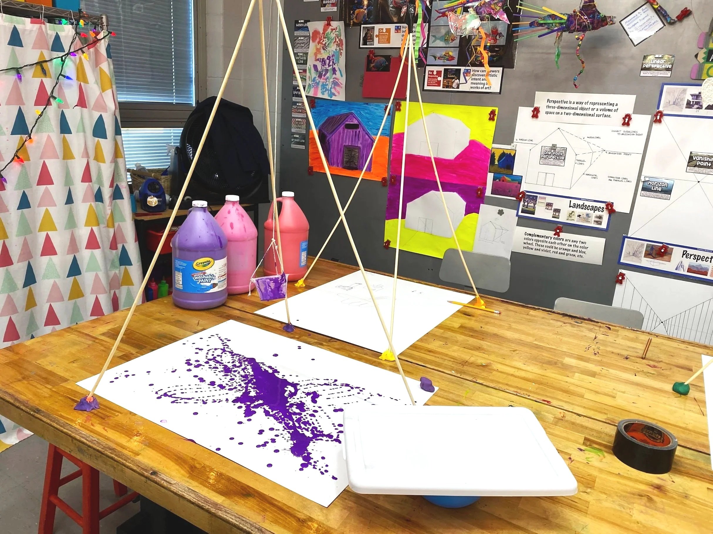 An art classroom workspace with a wooden table covered in purple paint splatter on paper, bottles of washable paint, and a geometric structure made of wooden sticks. A bulletin board in the background displays various art projects, posters, and descr