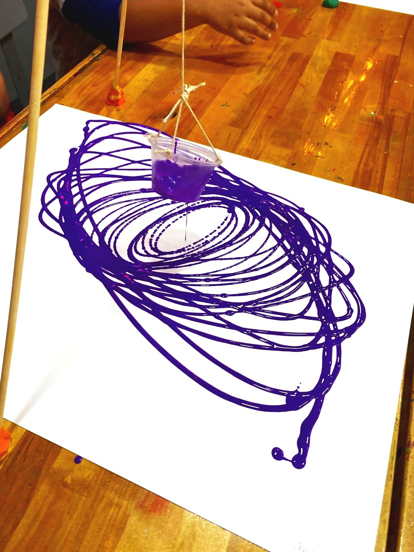 A science art project demonstrating liquid viscosity with purple paint, where a small plastic cup filled with purple paint is suspended over a white sheet of paper, creating purple swirl patterns on the paper.