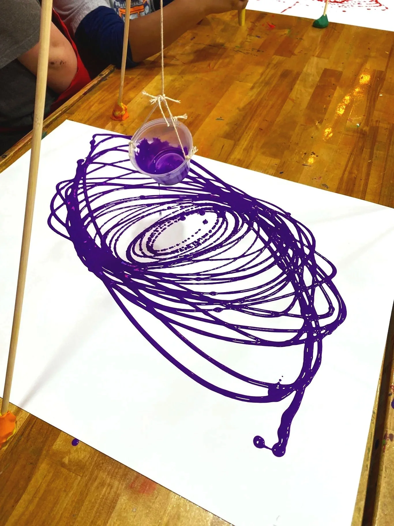 A colorful abstract art piece created with purple ink on white paper, with a small container of purple ink suspended above it by strings and sticks.