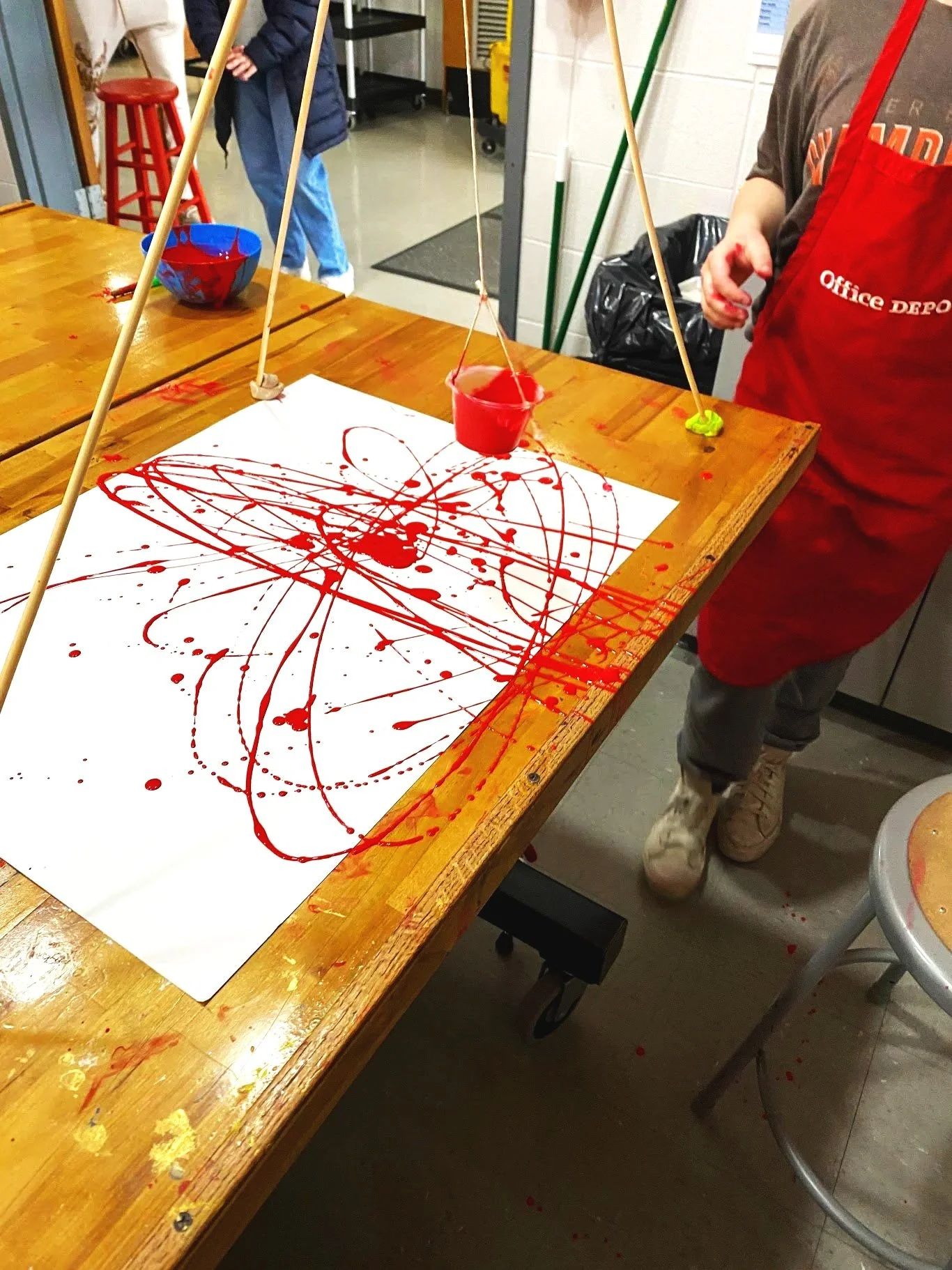 A table with an abstract painting created by dripping red paint onto a large white sheet of paper, with cups and sticks for paint pouring, in a casual art space.