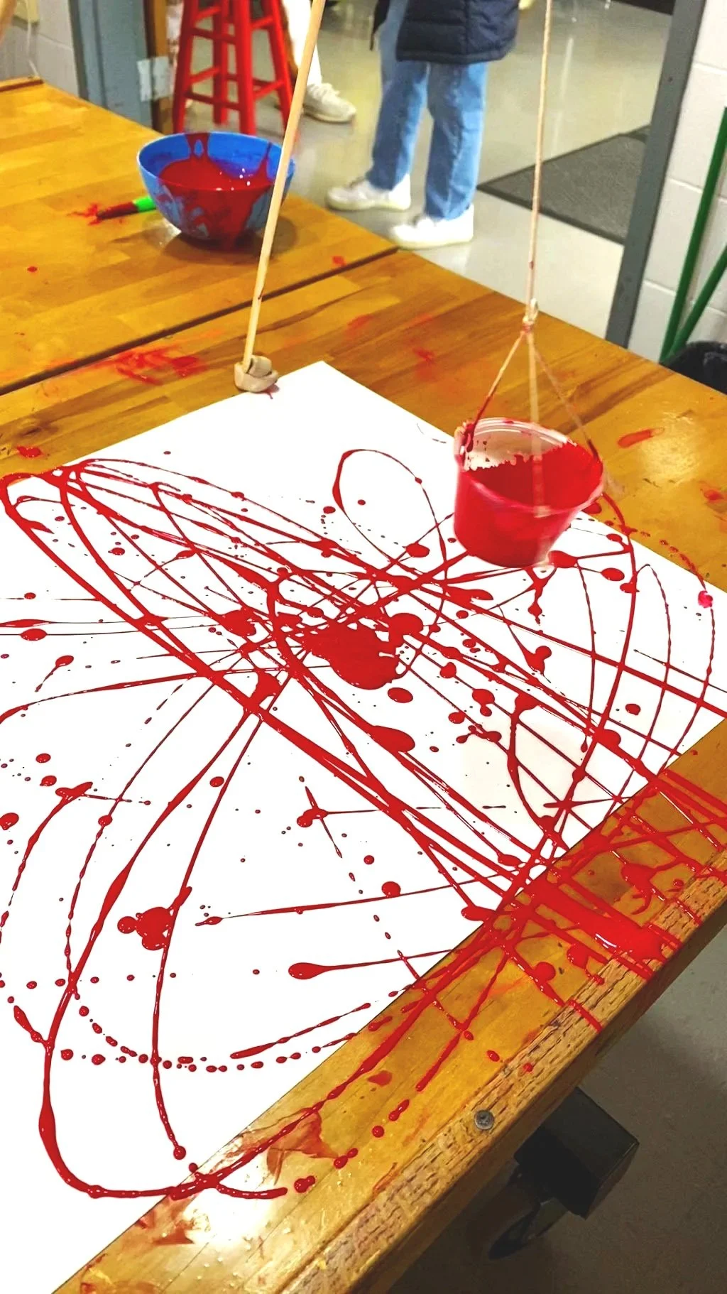 An art project using red paint, with cups hanging over a large sheet of white paper, which has red paint splattered and drizzled across it.