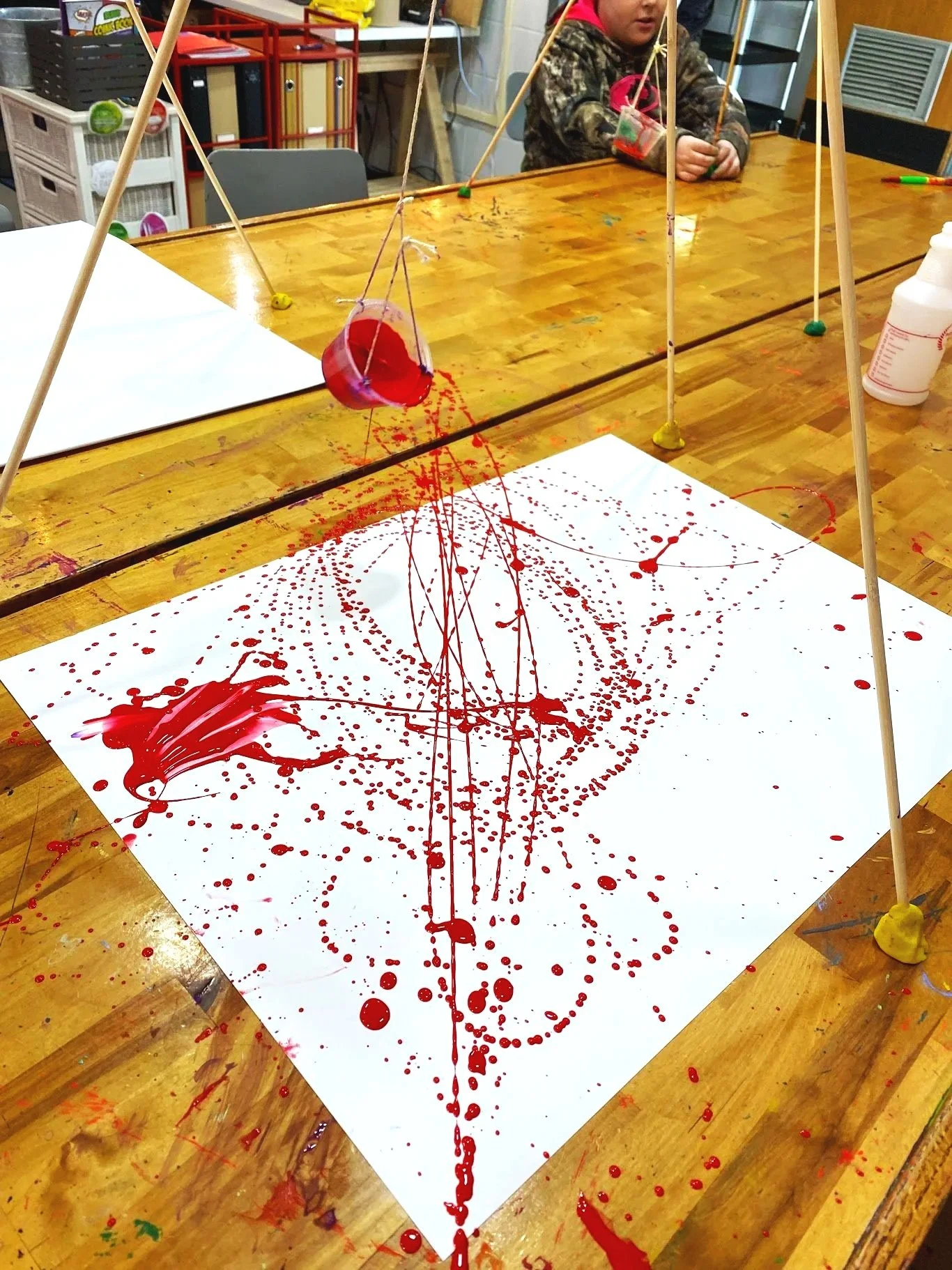 A science experiment setup with a paper on a table, splattered with red liquid. Several wooden sticks with strings are arranged to hold a small container filled with red liquid above the paper, creating splatters.