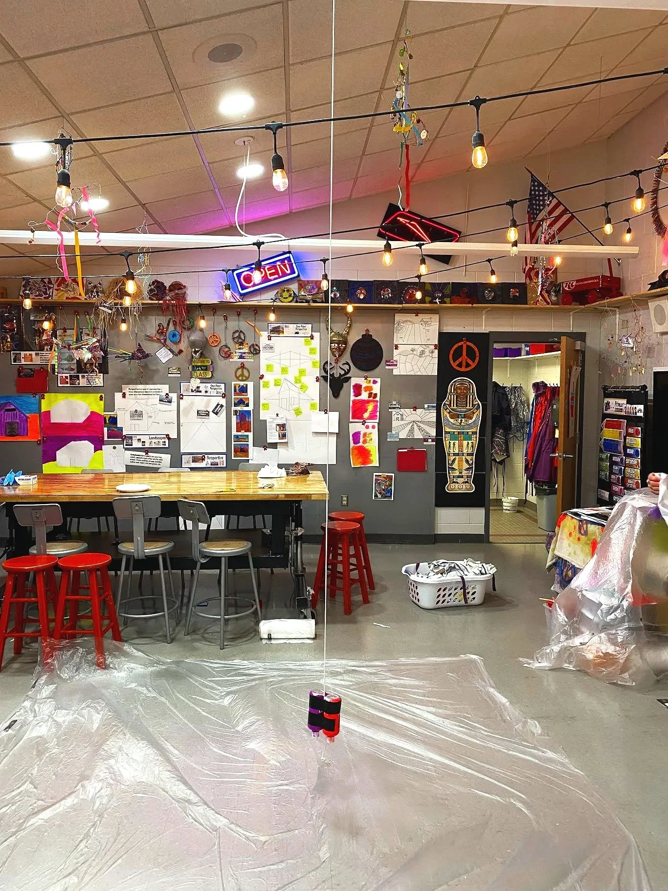 The image shows an art classroom or studio with a large mirror in the center, reflecting the room. There are string lights hanging from the ceiling, colorful artwork, and various art supplies and decorations on the walls. A large wooden table with ch