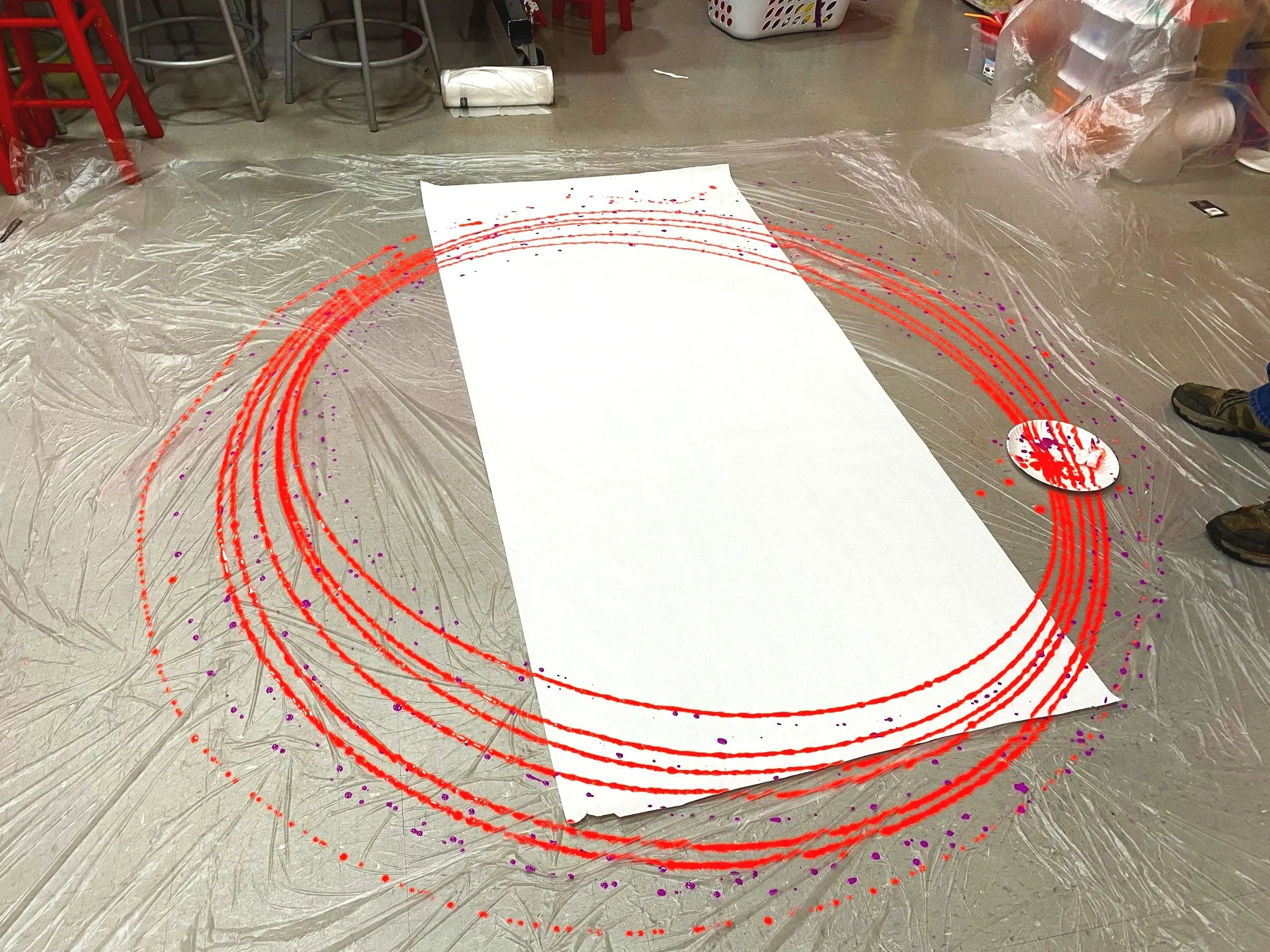 Large white canvas on the floor with red and purple spray paint circles and patterns. Multiple paints tied together created from a giant pendulum hanging from the ceiling. Art created by students with special needs.