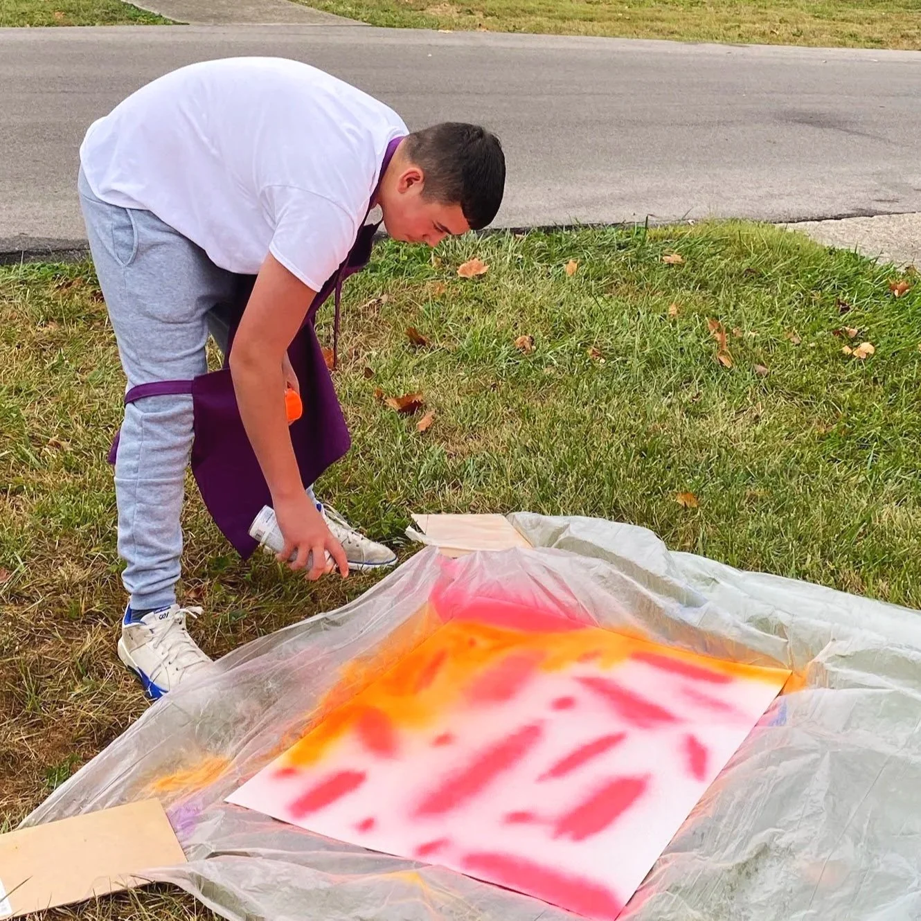 An adapted art project with a young man spray painting on a canvas outdoors, with paint colors red, orange, and yellow, on a plastic sheet on the grass.