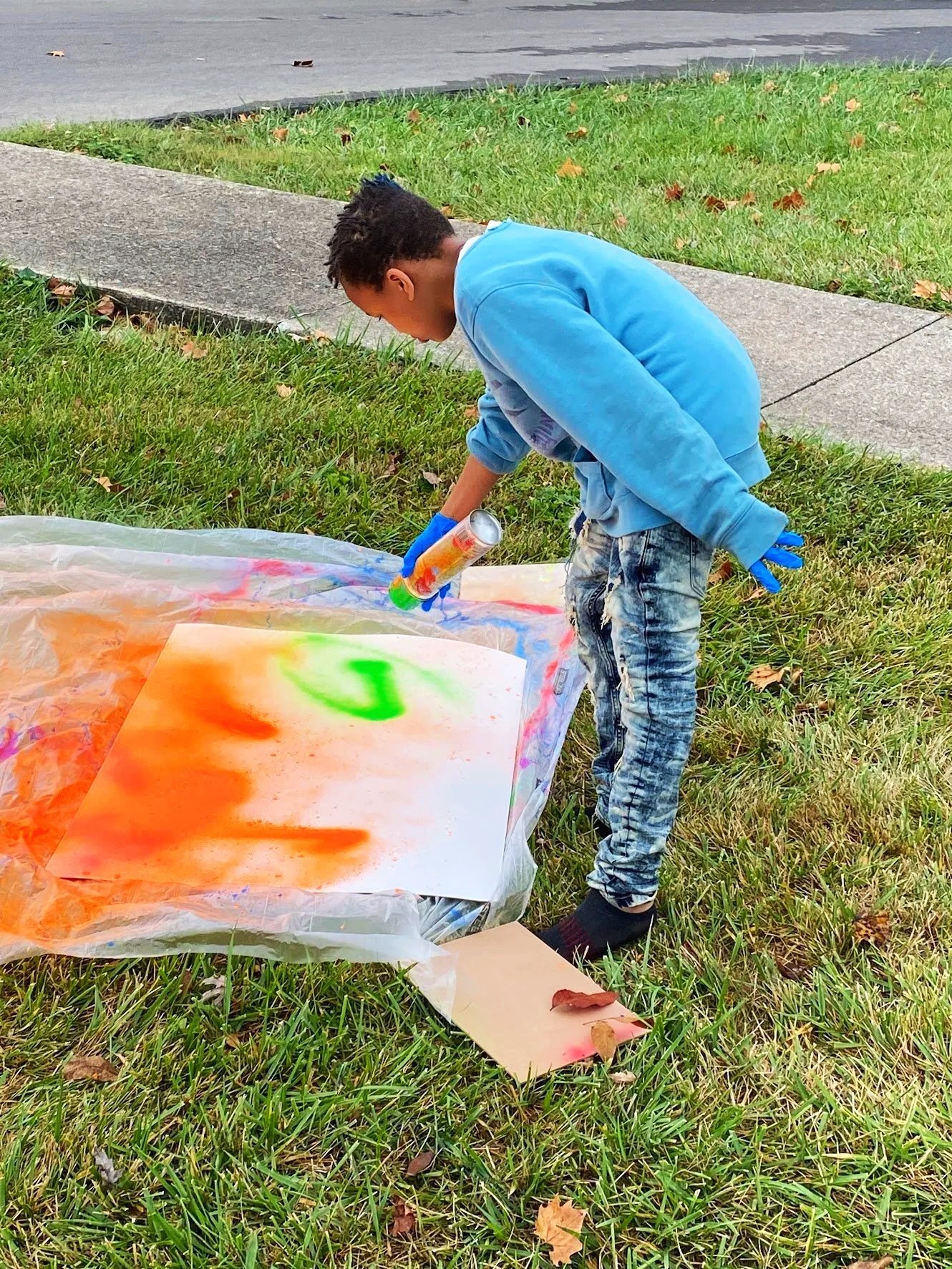 A young boy in a blue hoodie and ripped jeans is spray painting on paper placed on a plastic sheet outside, with grass and a sidewalk in the background.