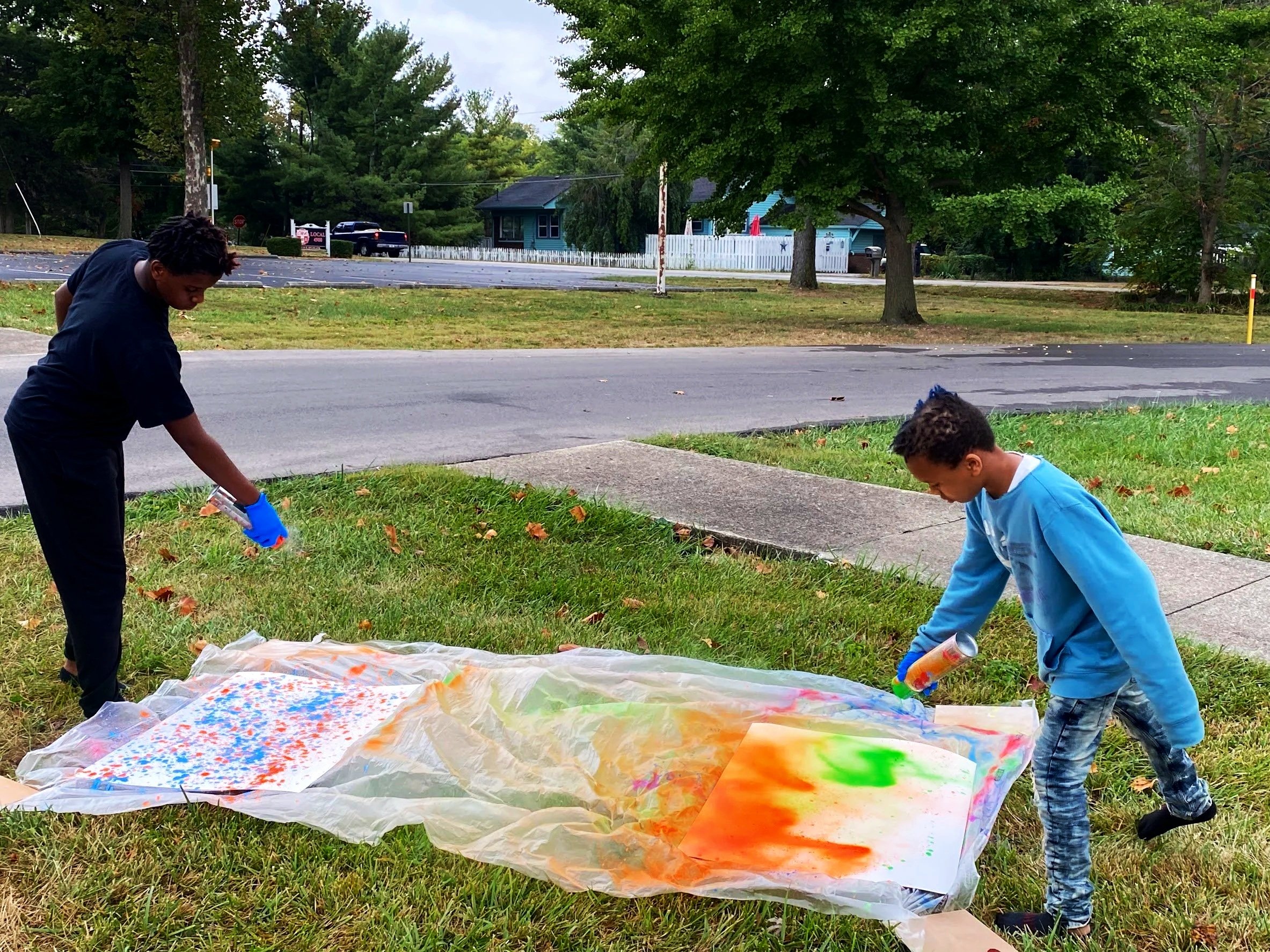 An a adapted art program with two children spray paint on large sheets of paper laid on a plastic sheet outdoors on grass, with trees, a street, and houses in the background.
