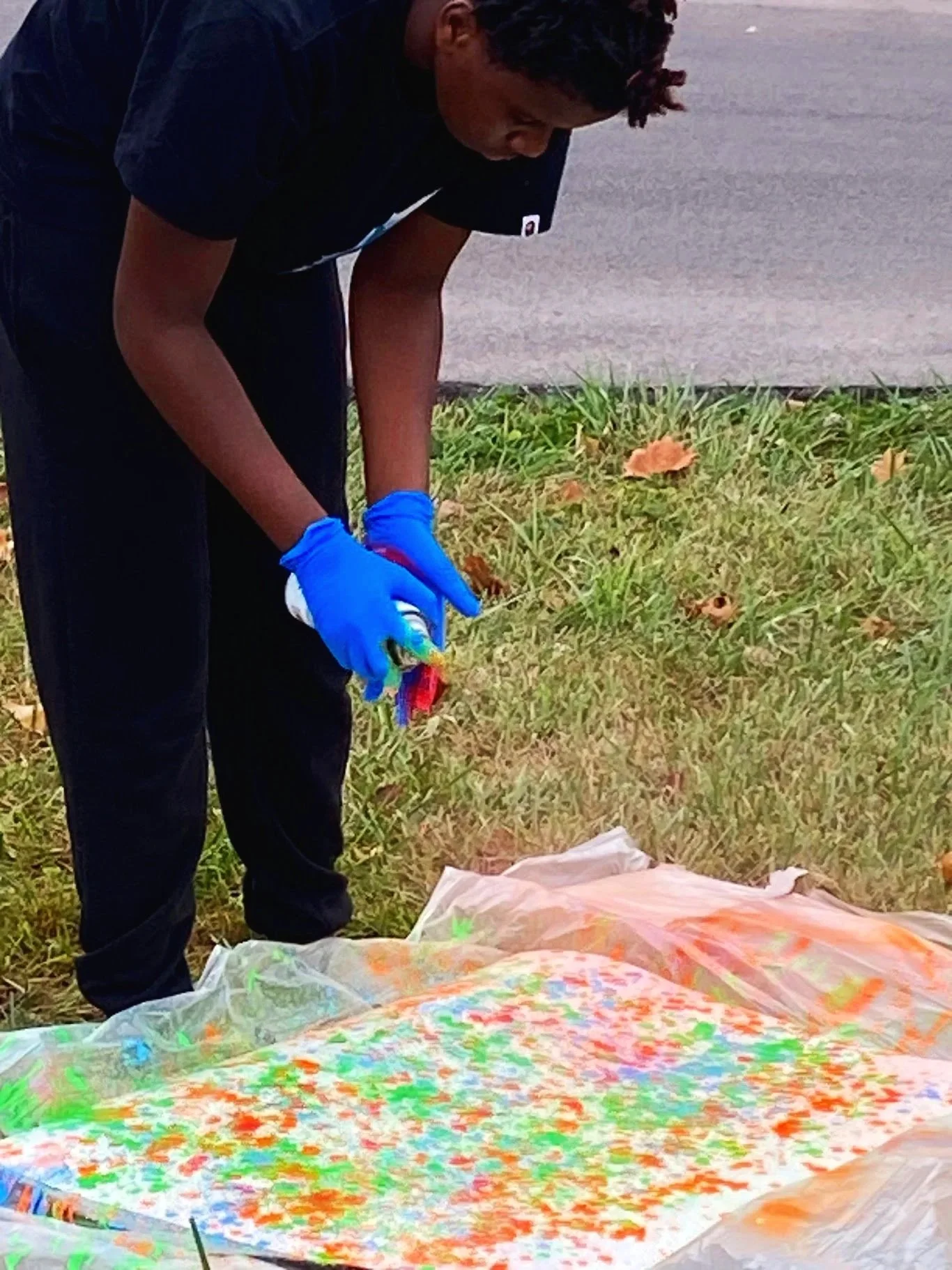 An art adapted project with a person wearing a black shirt and blue gloves is spray painting on a large plastic sheet laid on the grass, with multicolored paint splatters visible.