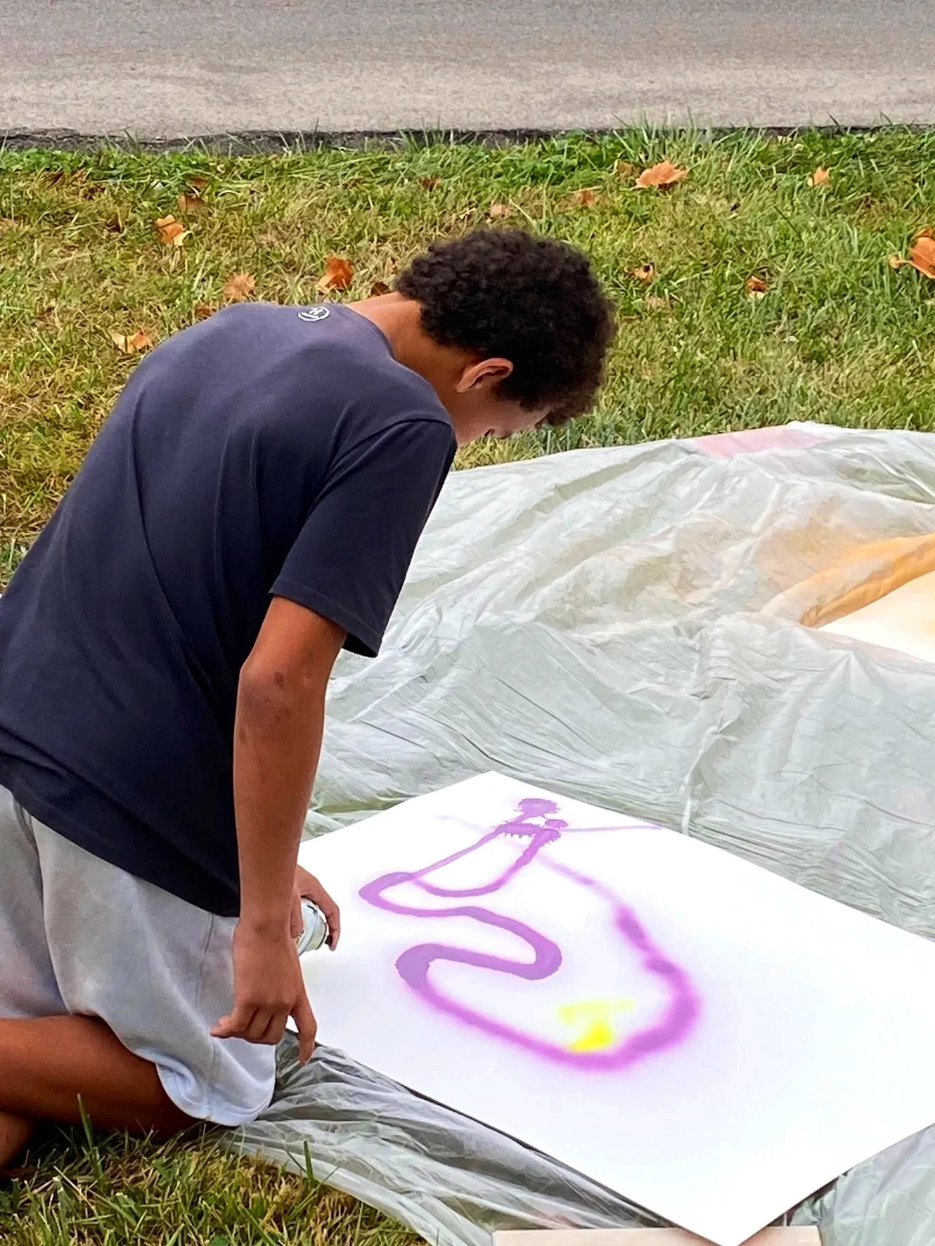 Person spray painting a design on a large white canvas outdoors on grass.