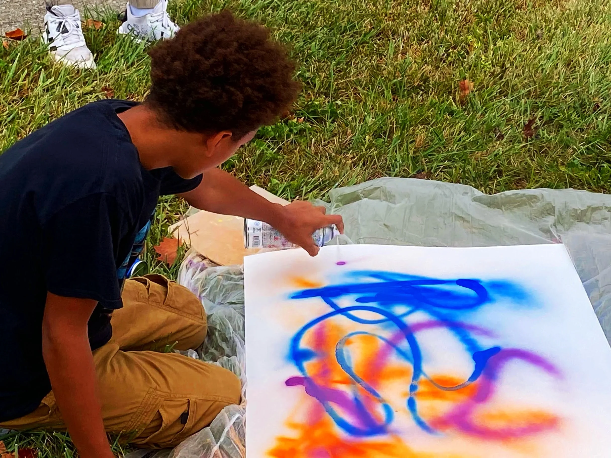 A young person kneeling on the grass, spray painting colorful abstract designs on a large sheet of paper outdoors.
