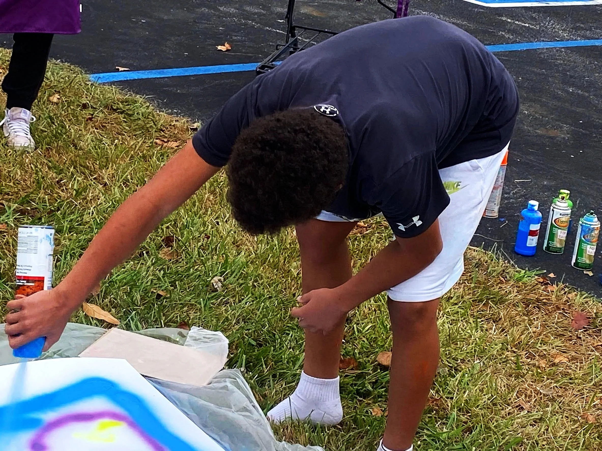 Young man in black T-shirt and white shorts crouches on grassy area painting on canvas outdoors.