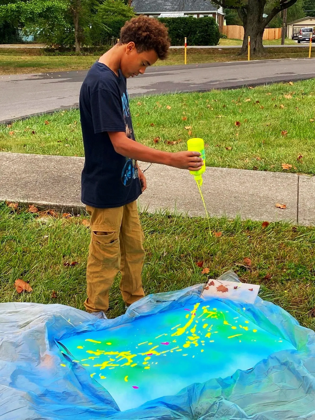 A young boy standing on grass outside, spray painting and creating graffiti art on a large sheet of paper on the ground.