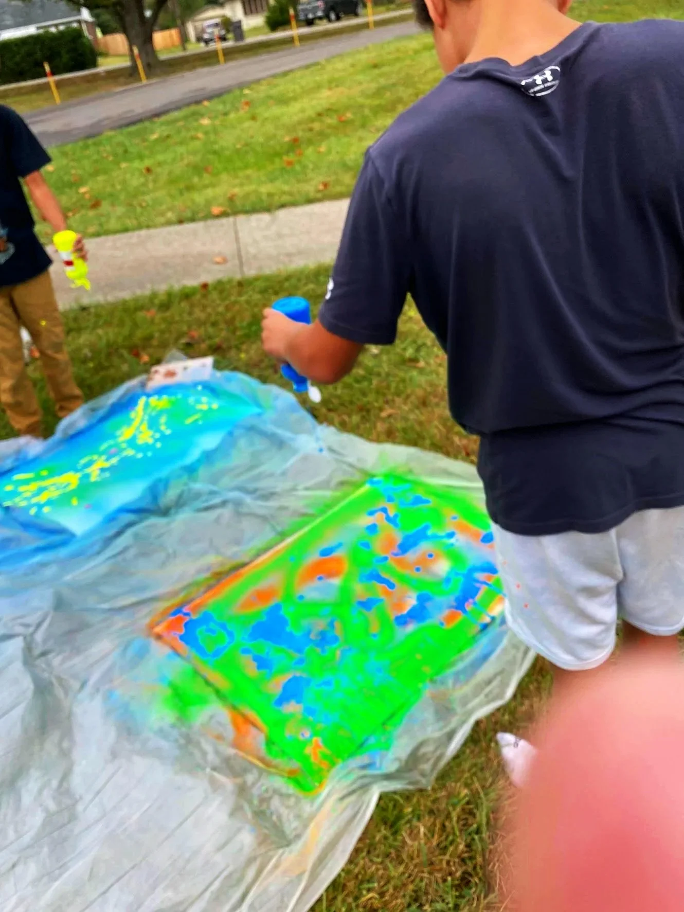 Students creating graffiti artwork with paint on a plastic-covered surface outdoors in a grassy area.