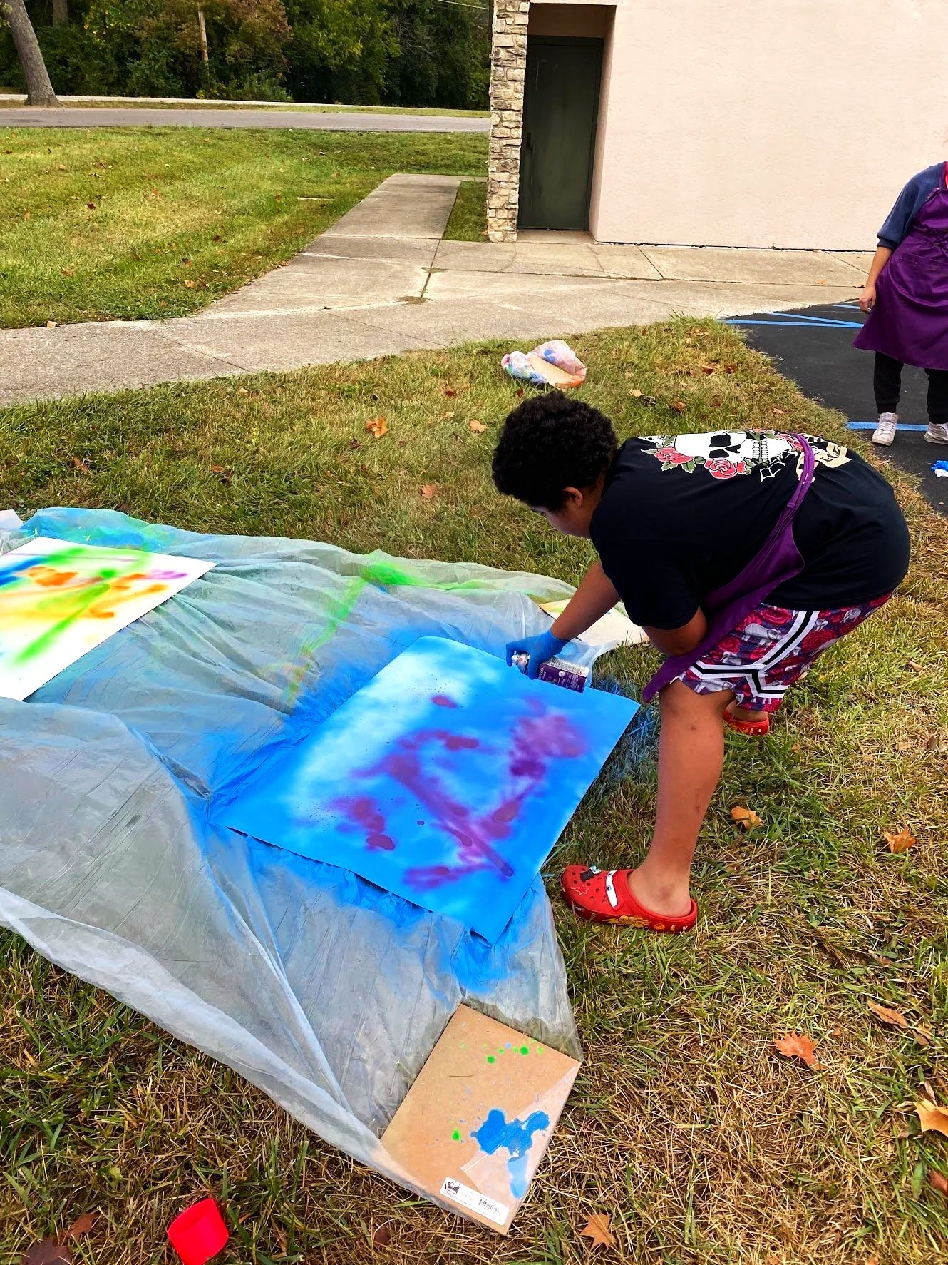 Child spray painting on a piece of paper on the ground outdoors, wearing a black T-shirt with floral print and red Crocs, with spray paint cans nearby.