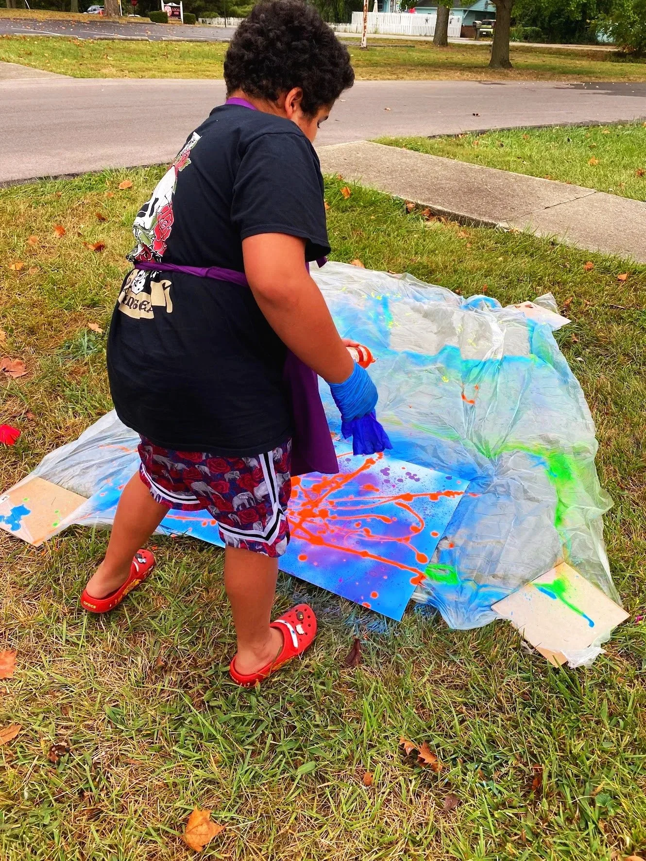 A boy with curly hair wearing a black t-shirt, floral shorts, red Crocs, and blue gloves is creating abstract splatter art on a canvas outdoors on a grassy area. The canvas is placed on a plastic sheet.