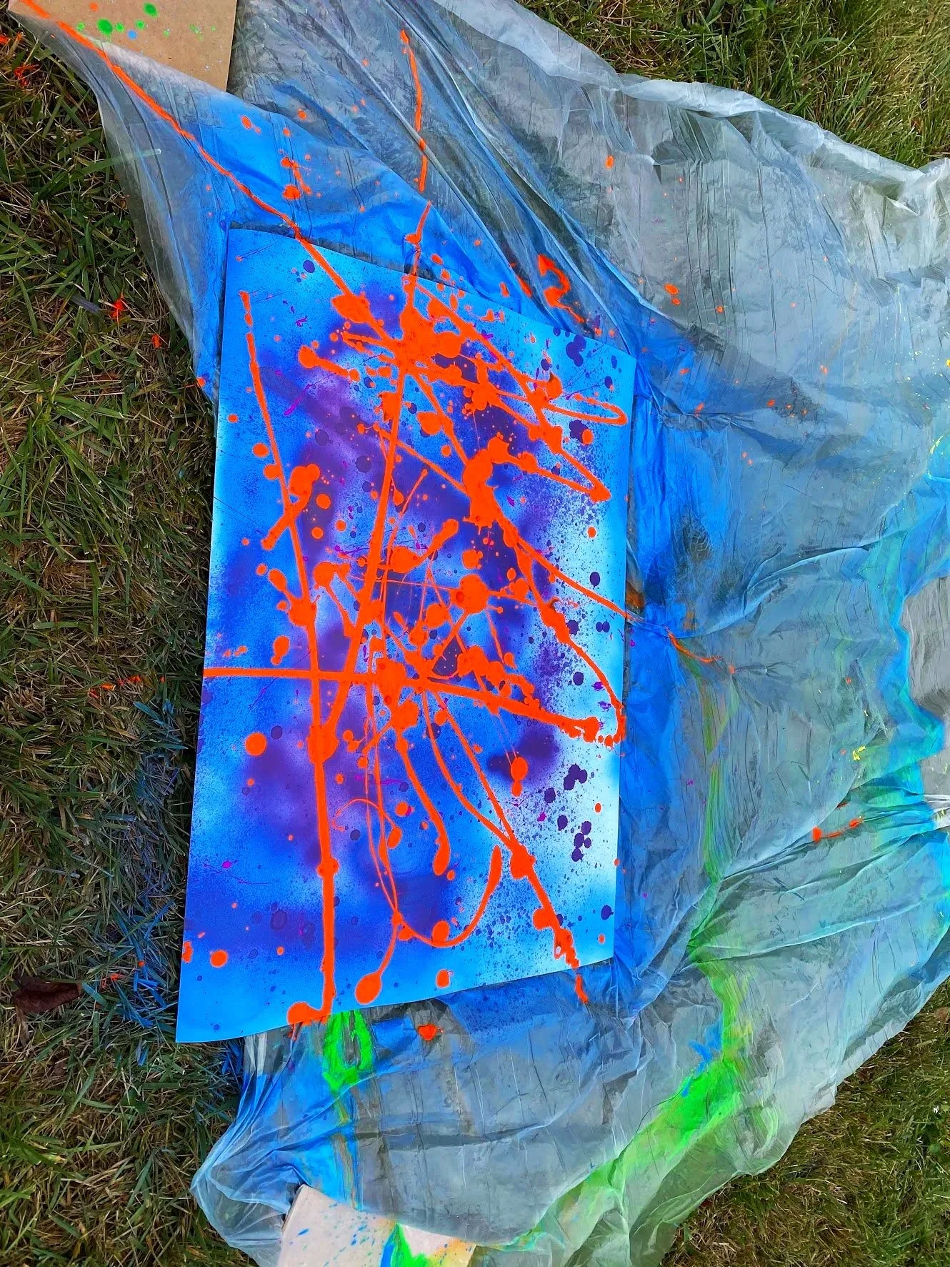 Abstract spray paint art on a blue canvas, with orange drips and splatters, on a grassy ground covered with a plastic sheet.