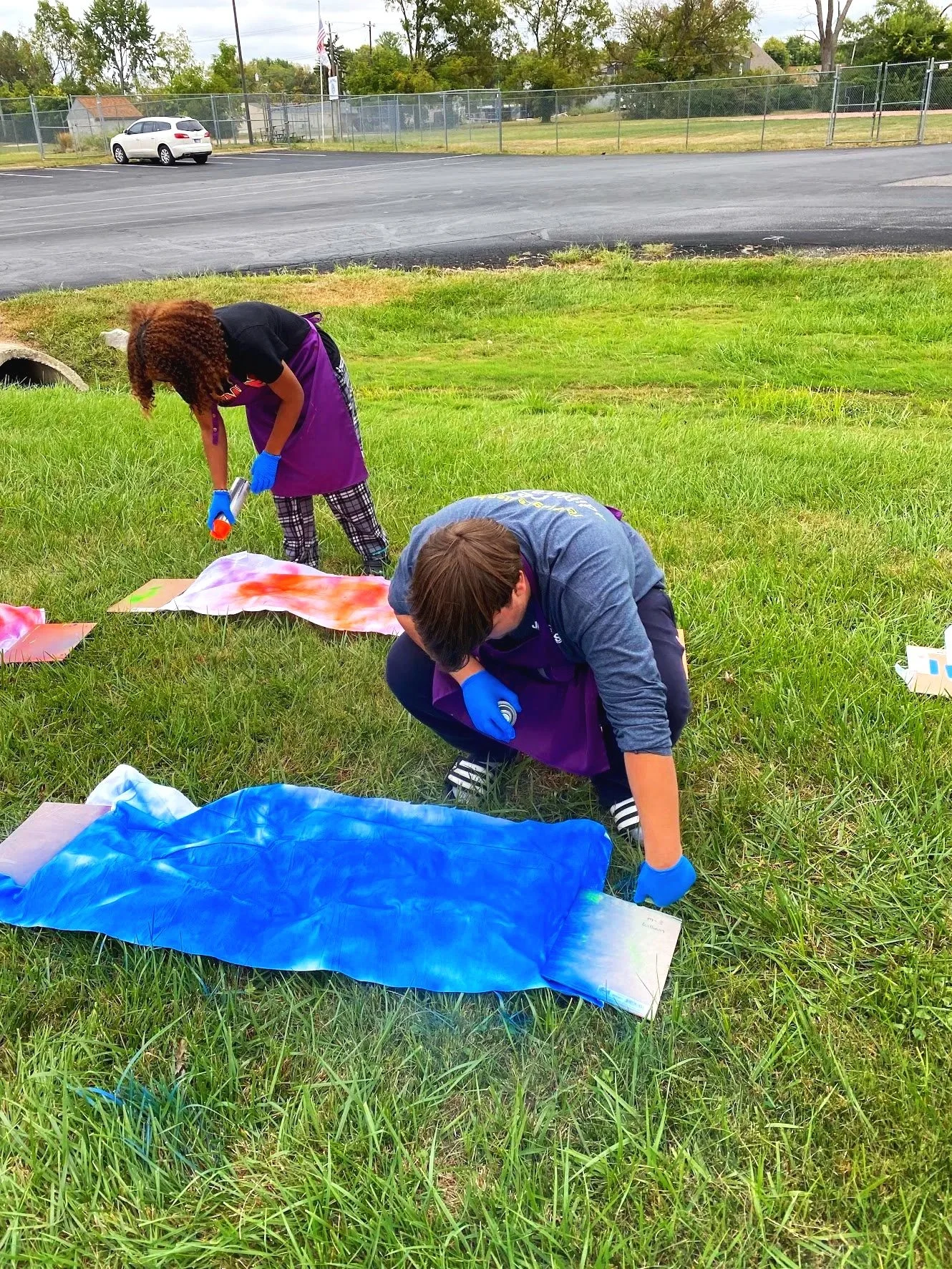 Two people crouch on grass painting large canvas-like objects outdoors. They wear blue gloves and purple aprons, with paint sheets on the ground nearby. The scene is set in an open grassy area next to a parking lot, with a chain-link fence and trees 