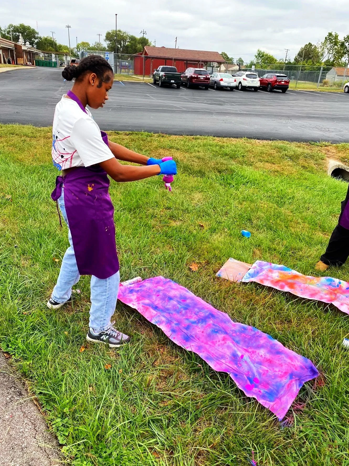A girl wearing a white T-shirt, purple apron, and gloves is spray painting a canvas on the grass outdoors during the daytime. There are multiple colorful painted canvases on the ground, and a parking lot with cars, a building, and cloudy sky in the b