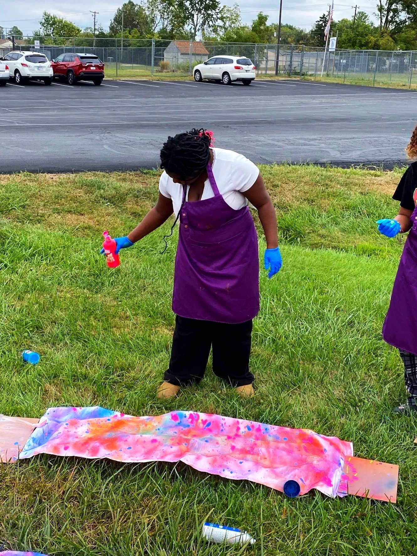 Woman with dreadlocks wearing a purple apron, white t-shirt, and blue gloves, spray painting on a large piece of fabric on grass near a parking lot.