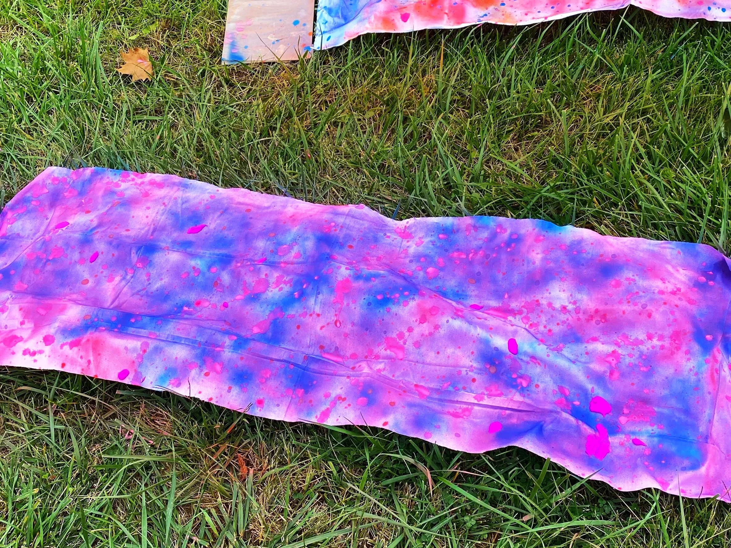 Large, colorful, tie-dye fabric with pink, purple, and blue splatters lying on green grass