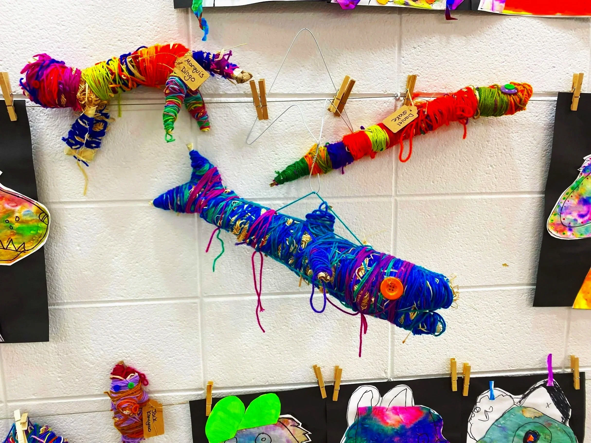 Colorful homemade yarn great white shark art project inspired by australian aboriginal Tjanpi artists hanging on a wire with clothespins, displayed on a wall.