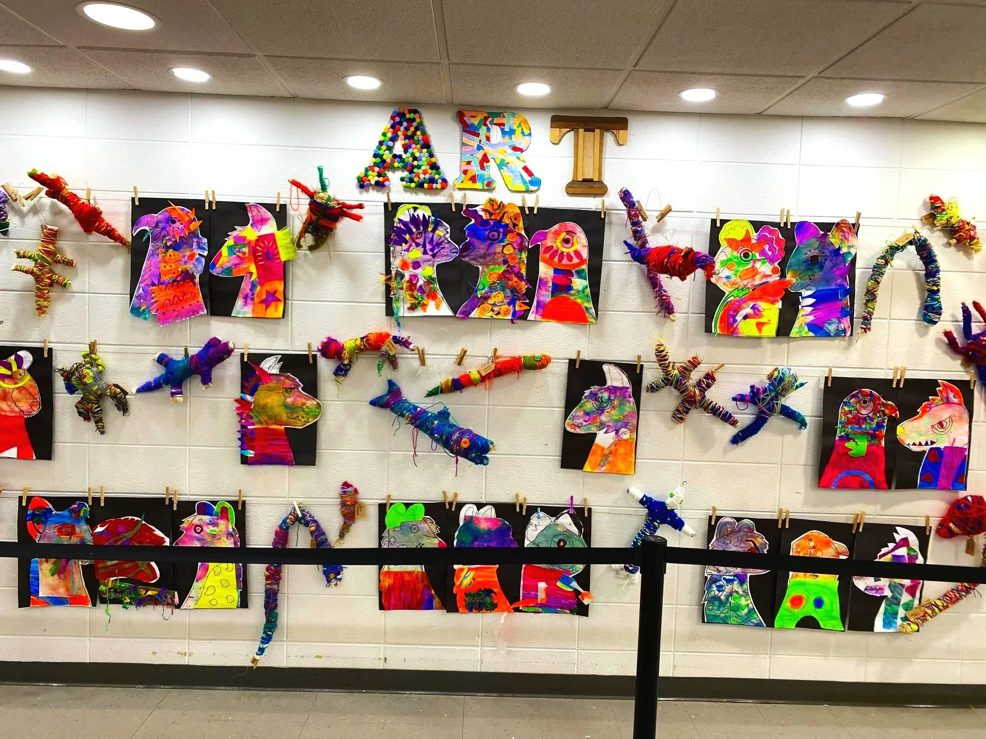 Colorful art display of unicorn and horse drawings with 3D plush and paper crafts on a white tiled wall with the letters ART hanging above.