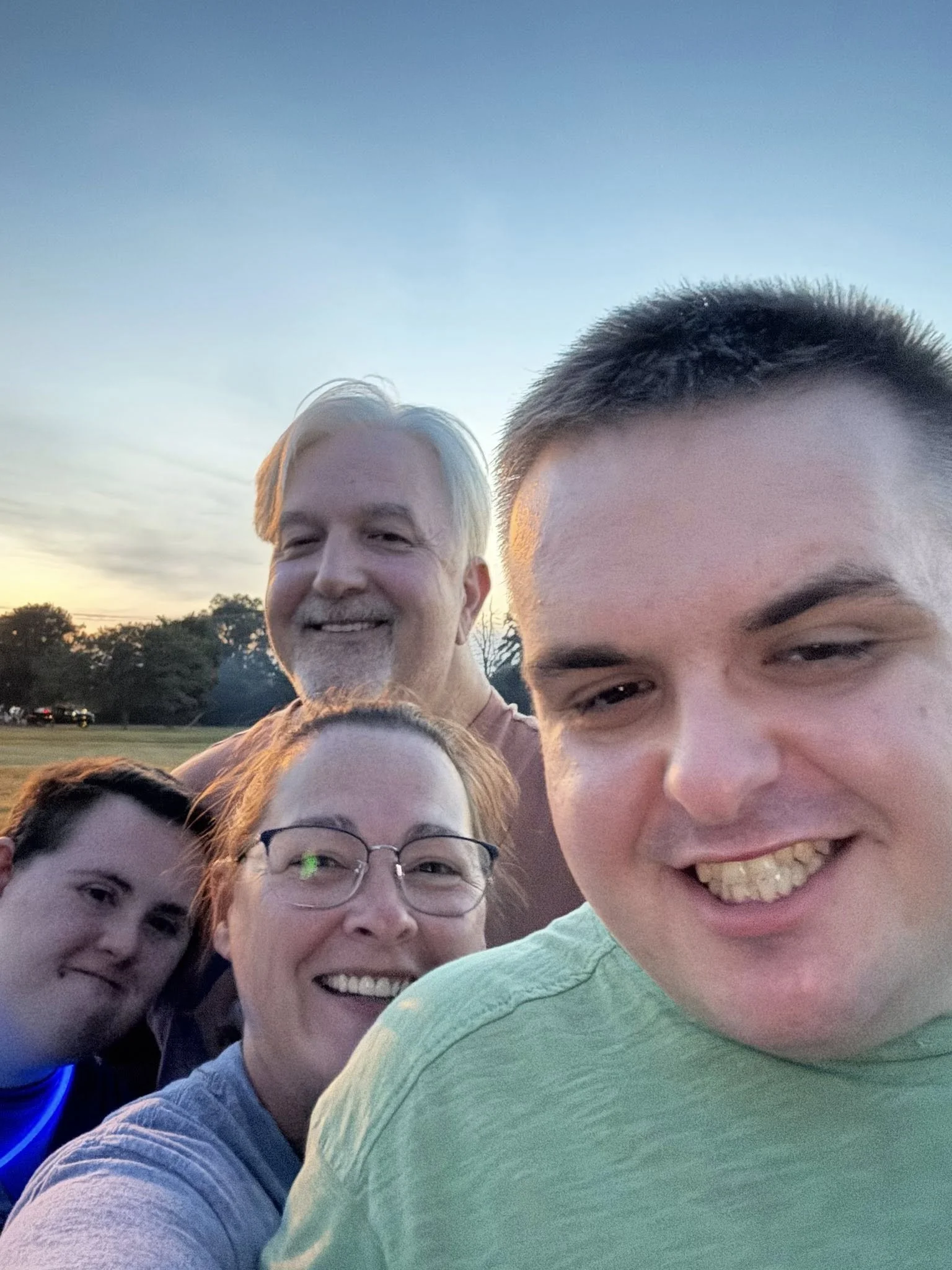 A group of smiling people, including children and adults, taking a selfie outdoors during sunset.
