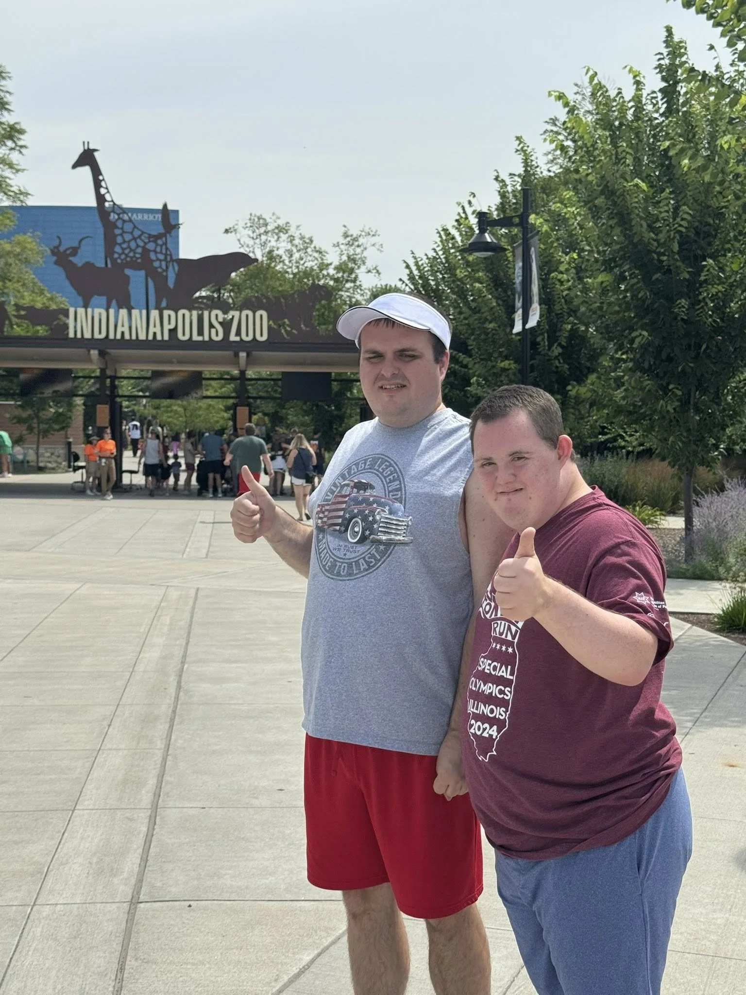 Two young men standing in front of the Indianapolis Zoo entrance, giving thumbs-up gestures, with trees and visitors in the background.