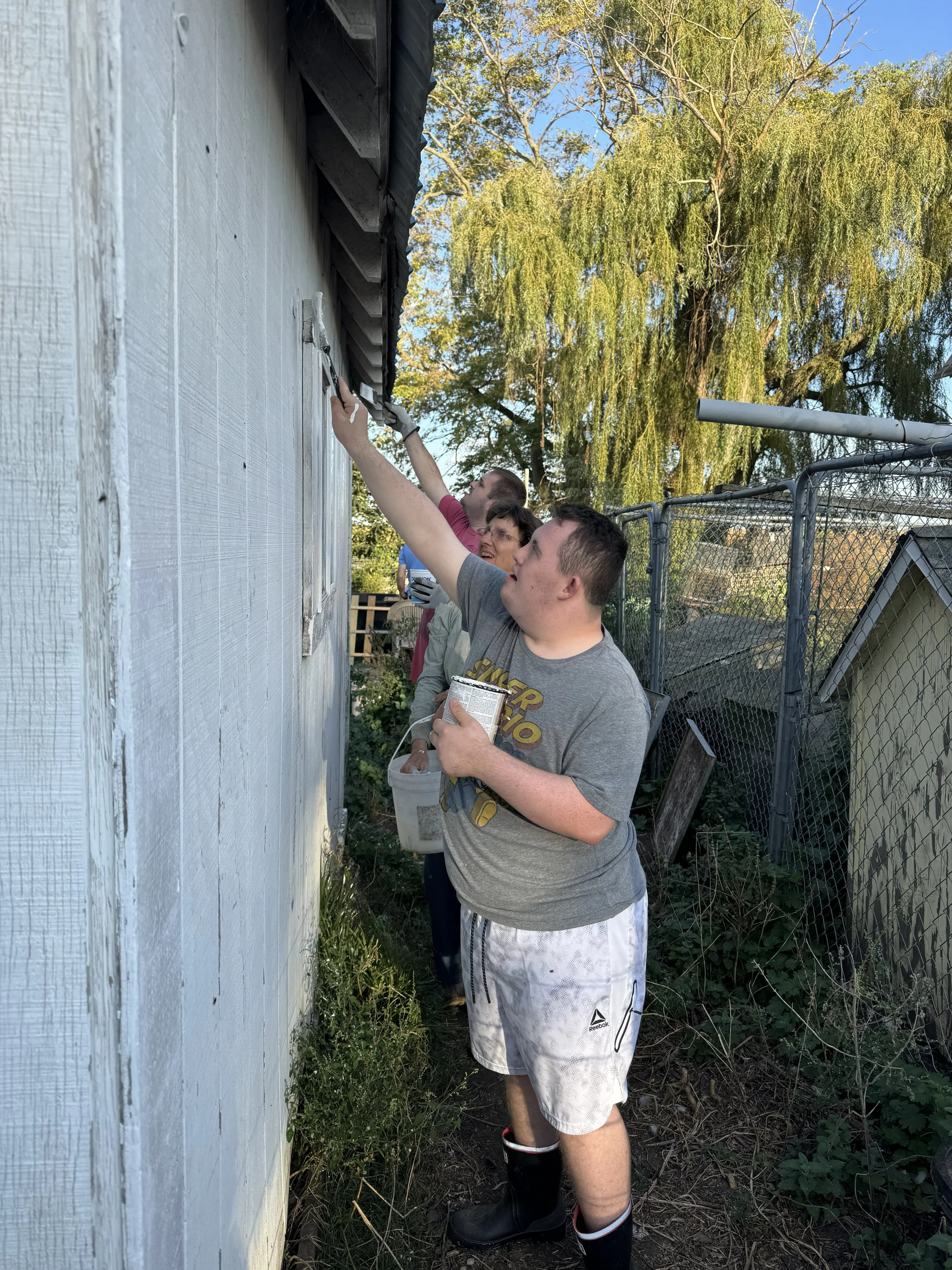 A young man in a gray t-shirt and white shorts paints a white wall with a small paintbrush while two women, one in a pink shirt and the other with glasses, observe and smile. They are outdoors near a house with a black roof, a chain-link fence, and a large green tree in the background.