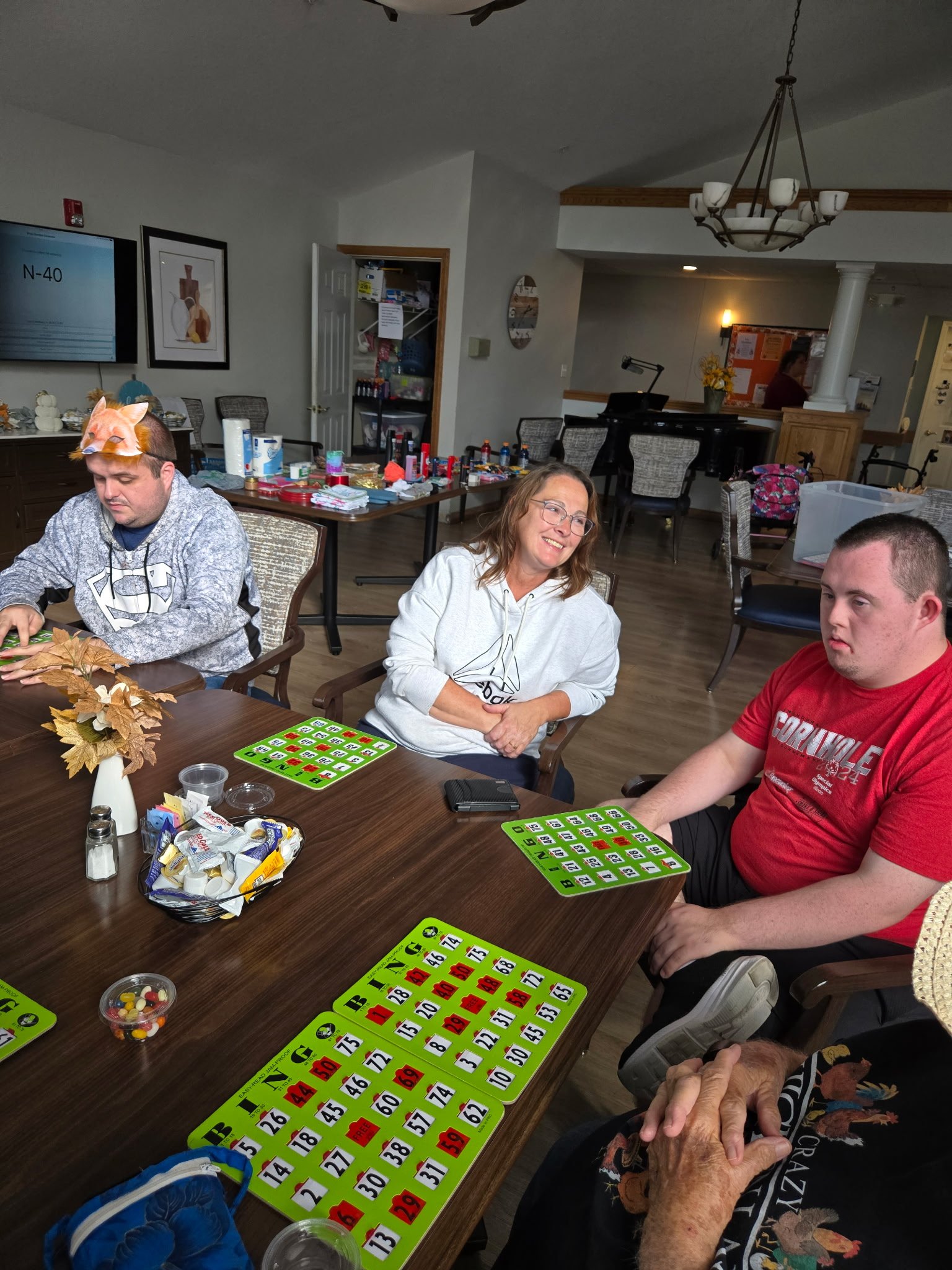 People sitting around a wooden table playing bingo, with bingo cards and markers, in a communal room.