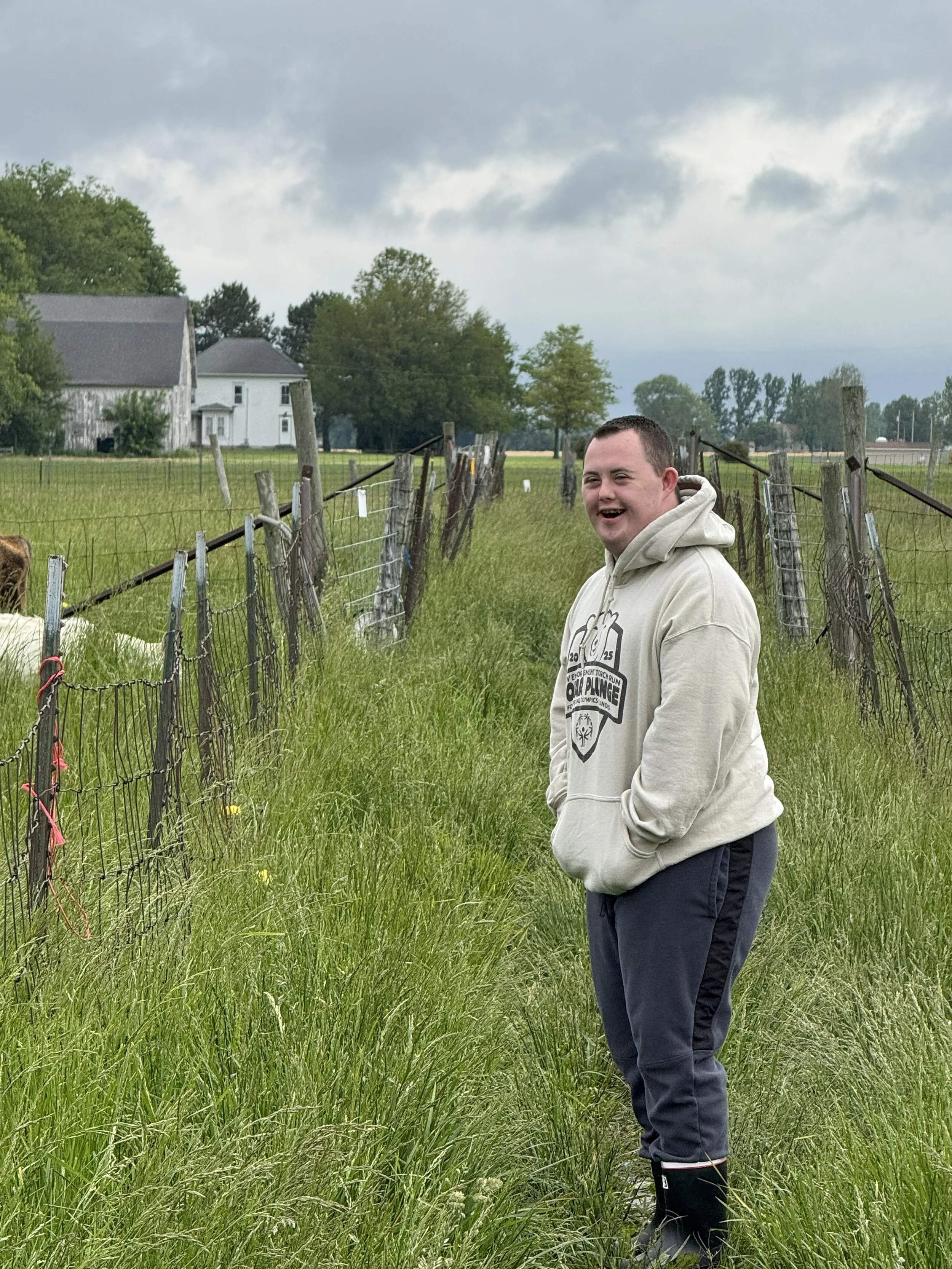 A young man with short hair wearing a beige hoodie, dark pants, and black rain boots, standing in a green grassy field next to a wire fence on a cloudy day, with trees and buildings in the background.