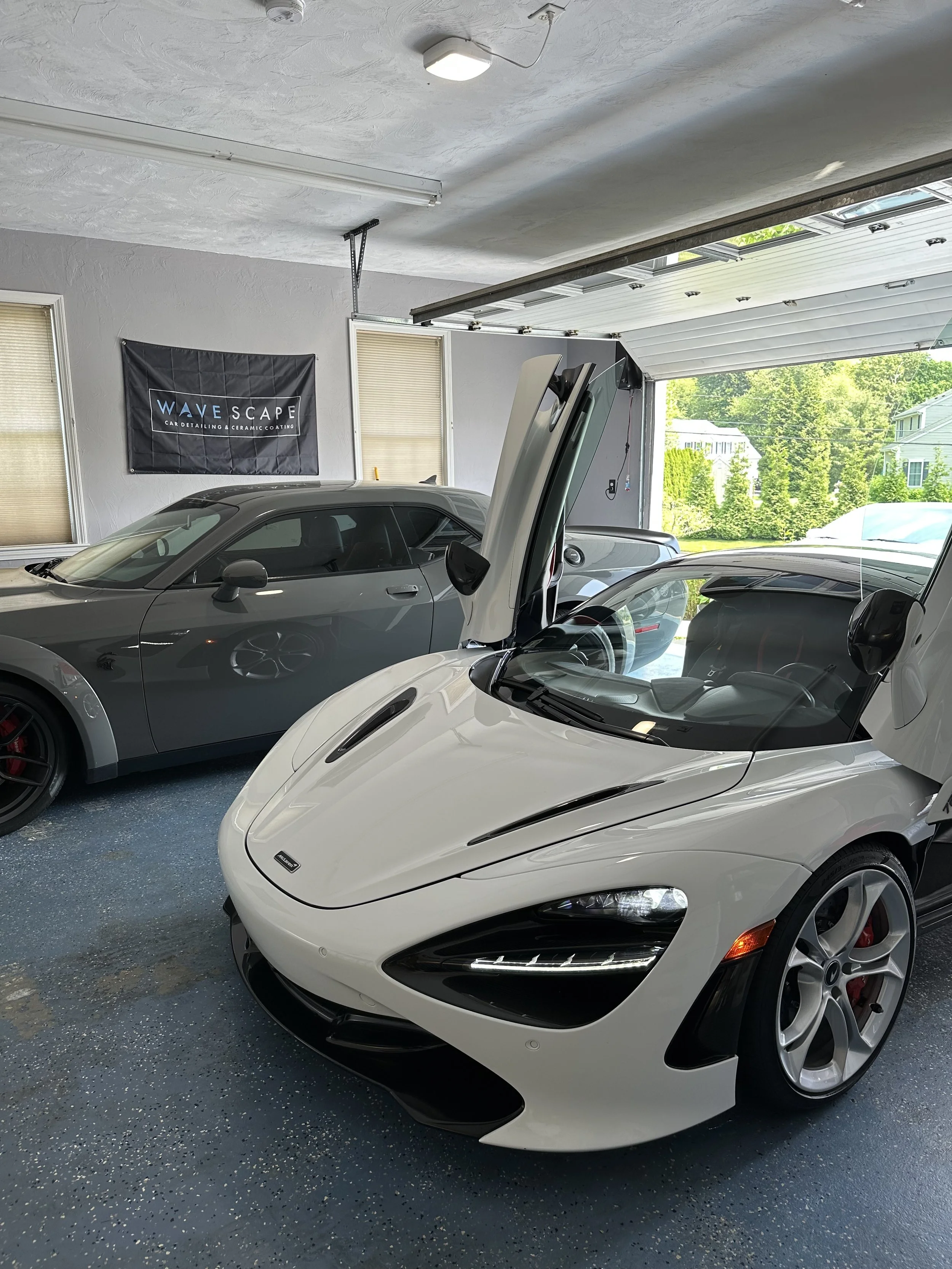 White sports car with butterfly doors open inside a garage, with another gray car parked nearby and a window showing a sunny outdoor scene.