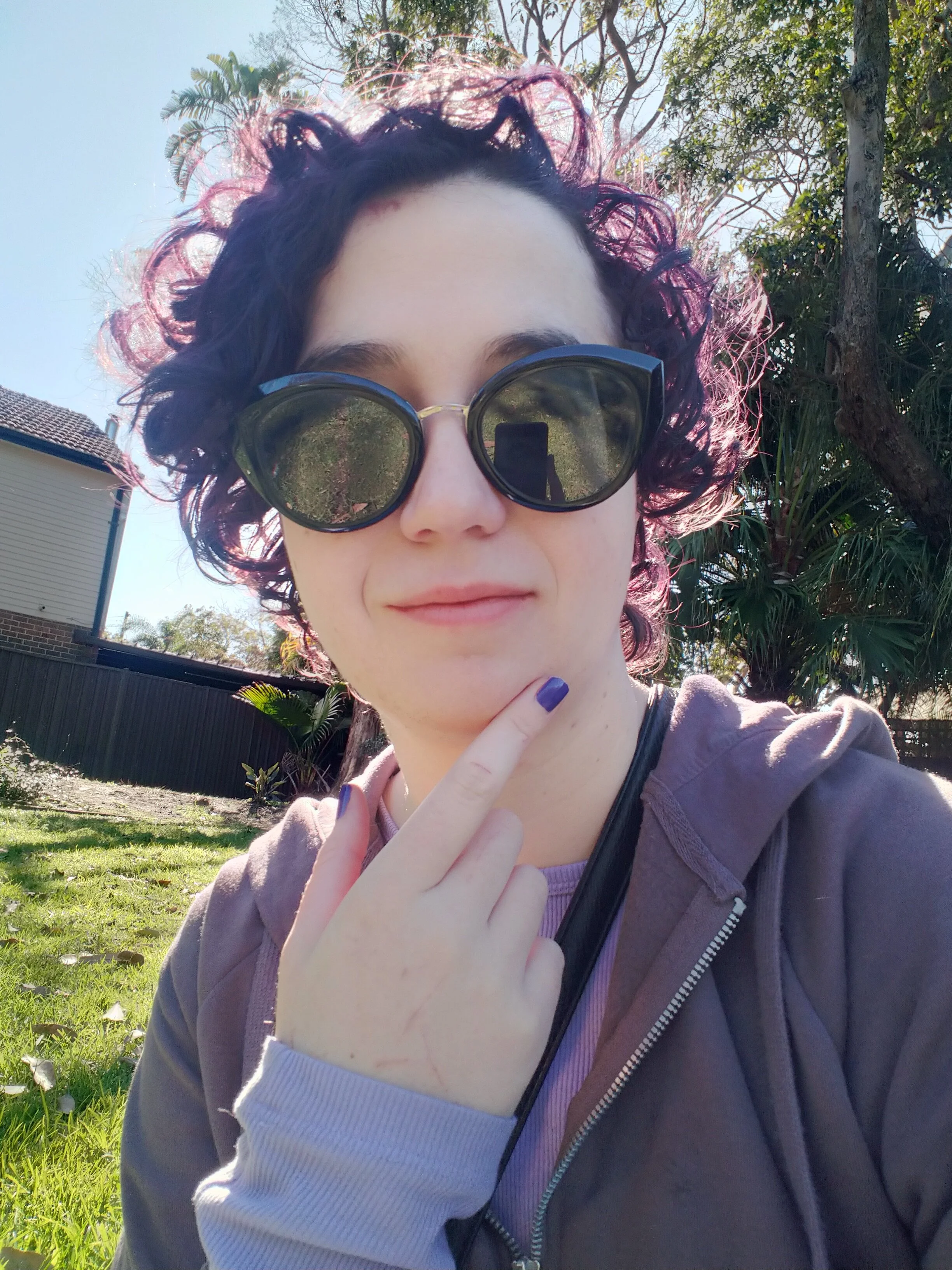 Person with short, curly, purple hair and large black sunglasses taking a selfie outdoors on a sunny day. Person has lavender nails and is wearing a purple hoodie over a shirt, with a black strap across their shoulder. Background includes green grass, trees, and a house.