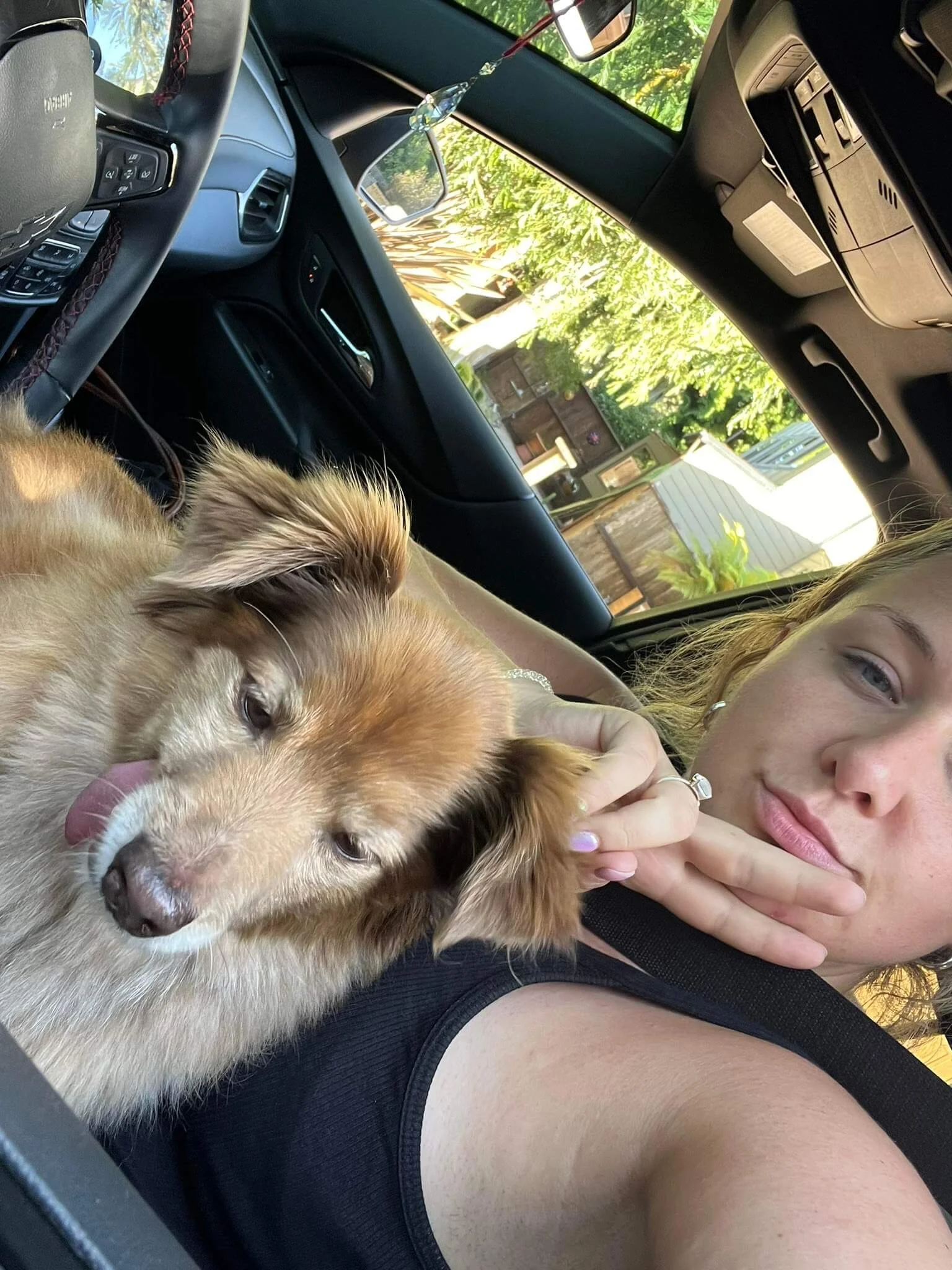 A woman lying in a car with a fluffy brown and white dog resting on her chest, both appear relaxed. The woman has light skin and blonde hair, and is wearing a black tank top. The dog has a pink tongue and looks content. Outside the car window, there are green trees and a garden with a shed.