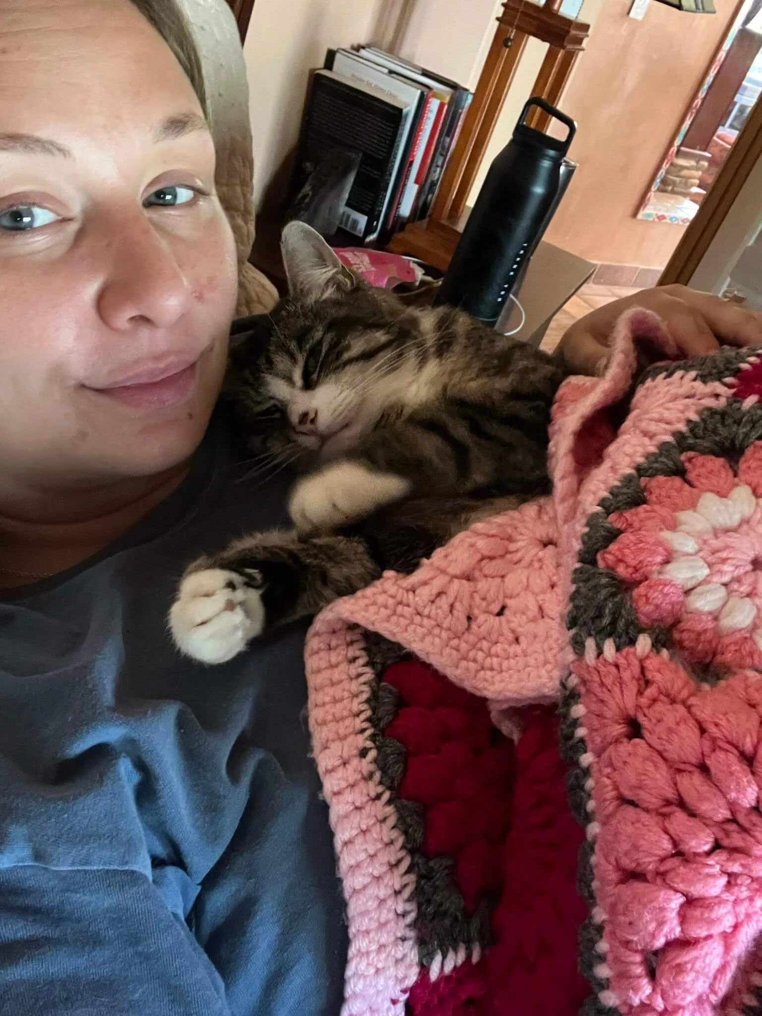 A person with blue eyes and a slight smile lying next to a sleeping tabby cat with striped fur, on a stuffed blanket. In the background, there are shelves with books and a black water bottle on a table.