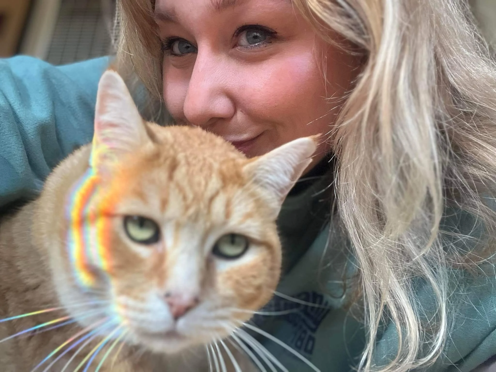 A woman close to a ginger cat with green eyes. The woman has blonde hair and is smiling, while the cat is looking at the camera during a pet sit.