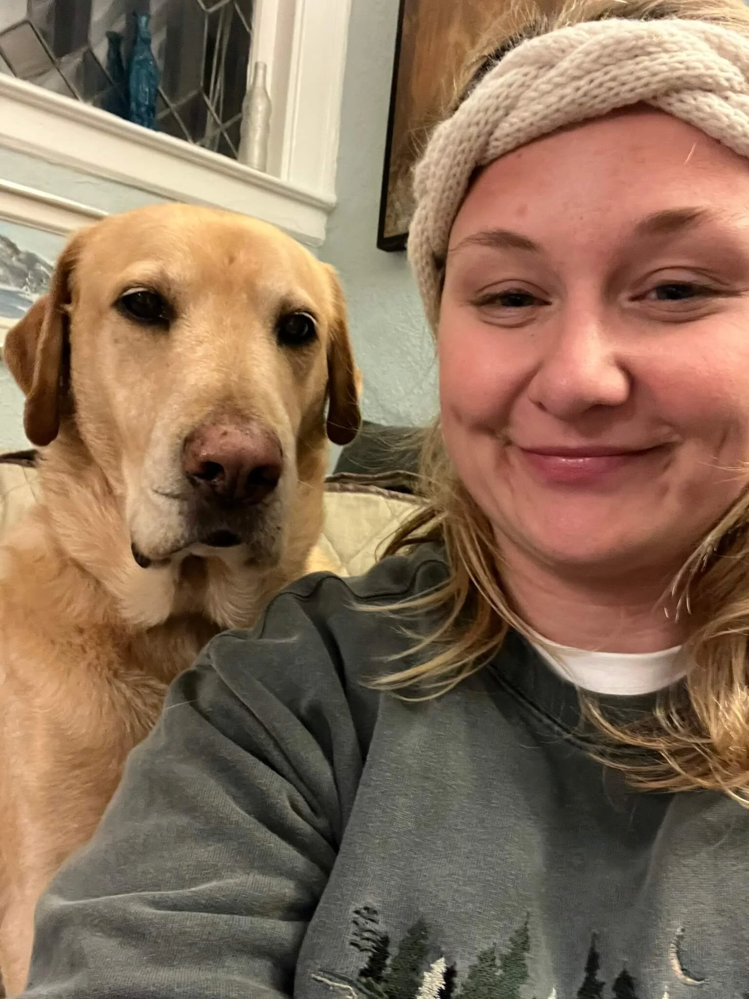 A woman with light skin and wearing a beige knitted headband smiling for a selfie next to her golden-colored dog.
