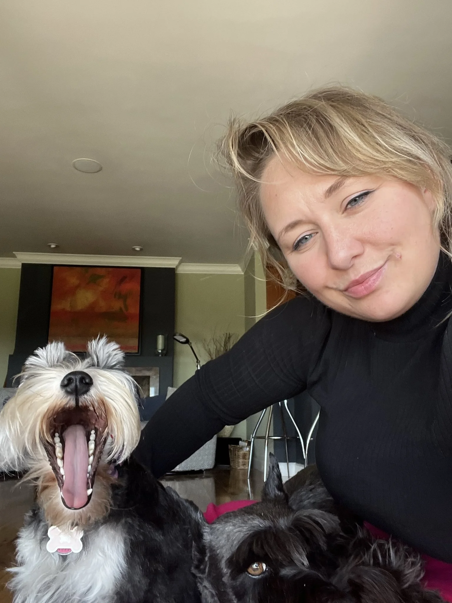 A woman smiling next to a yawning dog, both indoors with a green wall and artwork in the background.