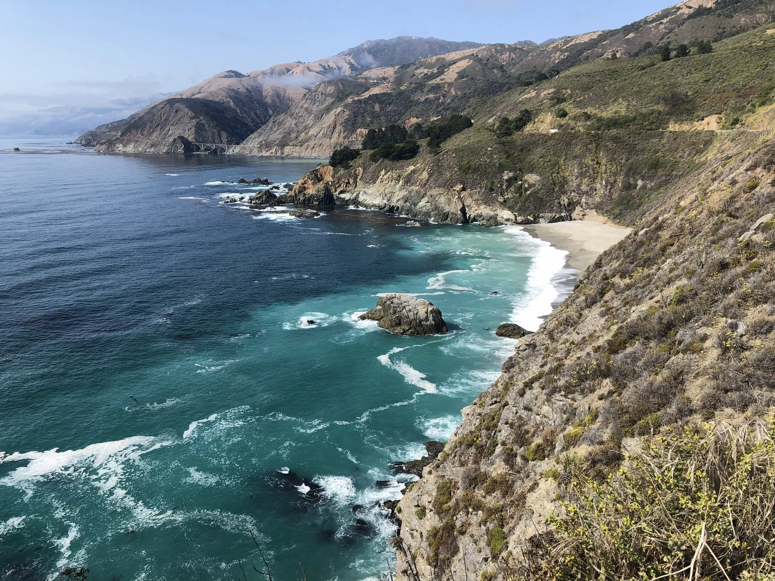 Scenic view of rugged cliffs and hills along a coastline, with ocean waves crashing against rocks and a bridge visible in the distance.