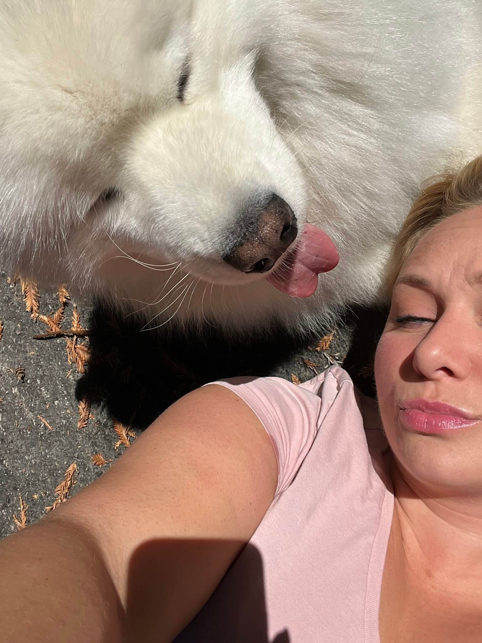 A person taking a selfie outdoors with a white dog licking their face. The person is wearing a light pink shirt, and the dog has white fur with a dark nose and black eyes. The ground is covered with small dried leaves. in-home pet sitting