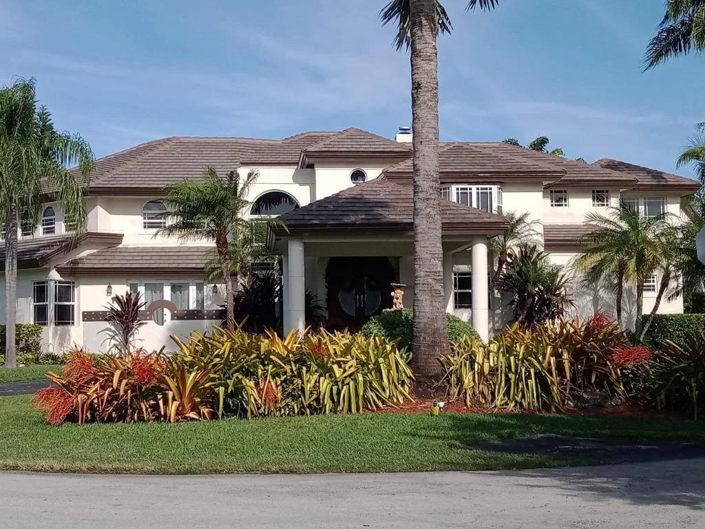 A large, two-story house with a brown tiled roof, white walls, and multiple windows, surrounded by palm trees and tropical plants in a well-maintained garden.