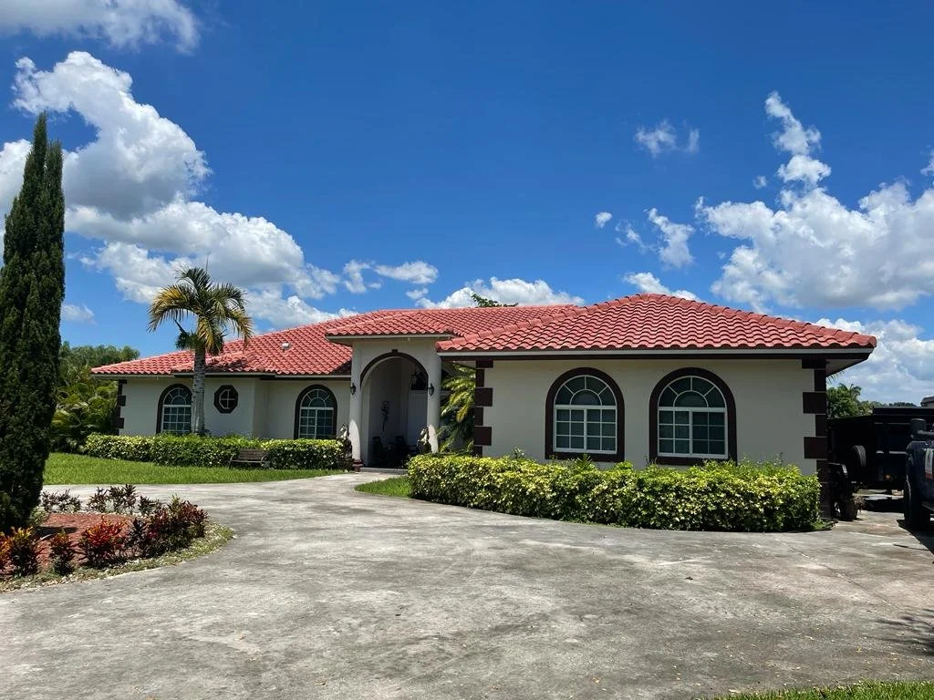 Single-story house with white walls, red tile roof, arched windows, surrounded by greenery and a paved driveway, under partly cloudy sky.