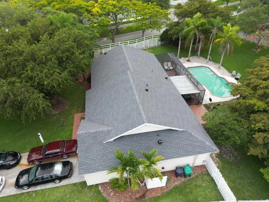 Aerial view of a suburban house with a gray roof, surrounding green trees, a small driveway with parked cars, and a backyard with a pool and palm trees.