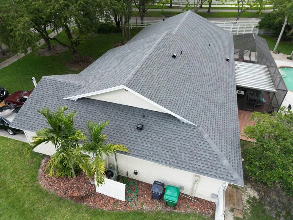 Aerial view of a house with a gray shingle roof, white exterior walls, a palm tree, three garbage bins, and a backyard with a screened pool enclosure and green lawn.