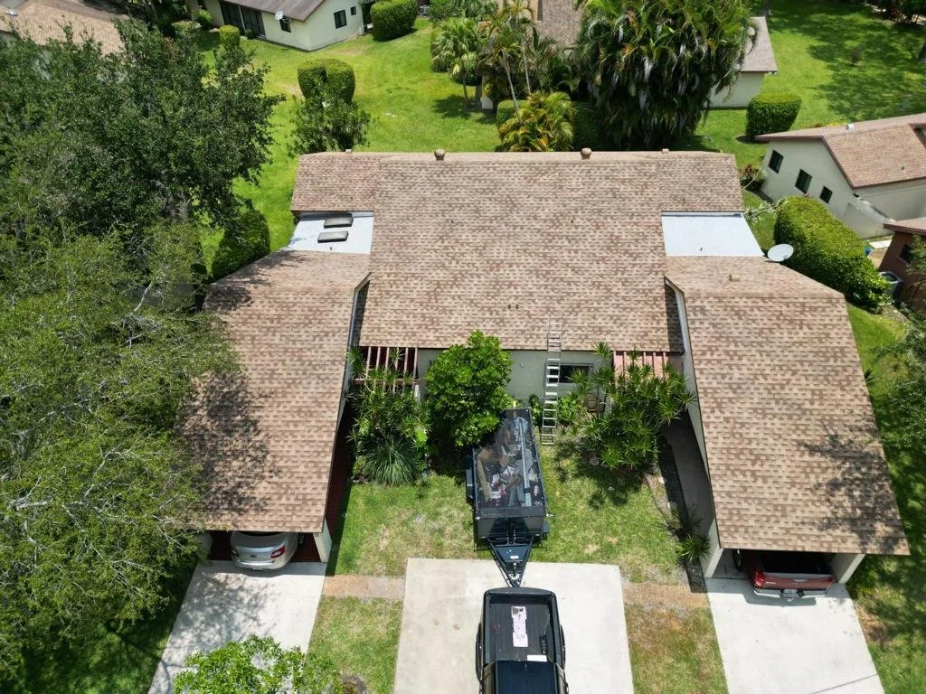 An aerial view of a house with a brown shingle roof, surrounded by green trees and yard. Two covered parking areas are on either side of a concrete driveway, with a trailer and a vehicle parked nearby. Ladders and tools are visible in the backyard, indicating ongoing work.